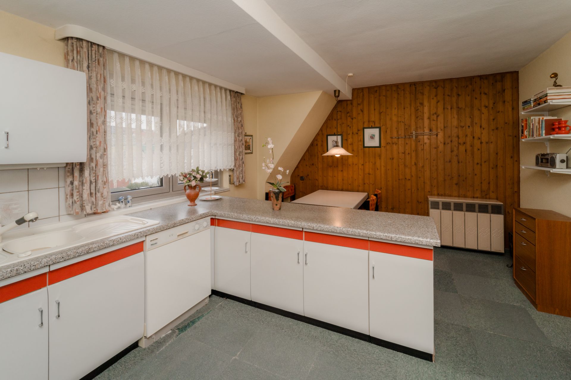 Küche mit weiß-roten Schränken, großem Fenster und angrenzendem Essbereich mit Holzwand.