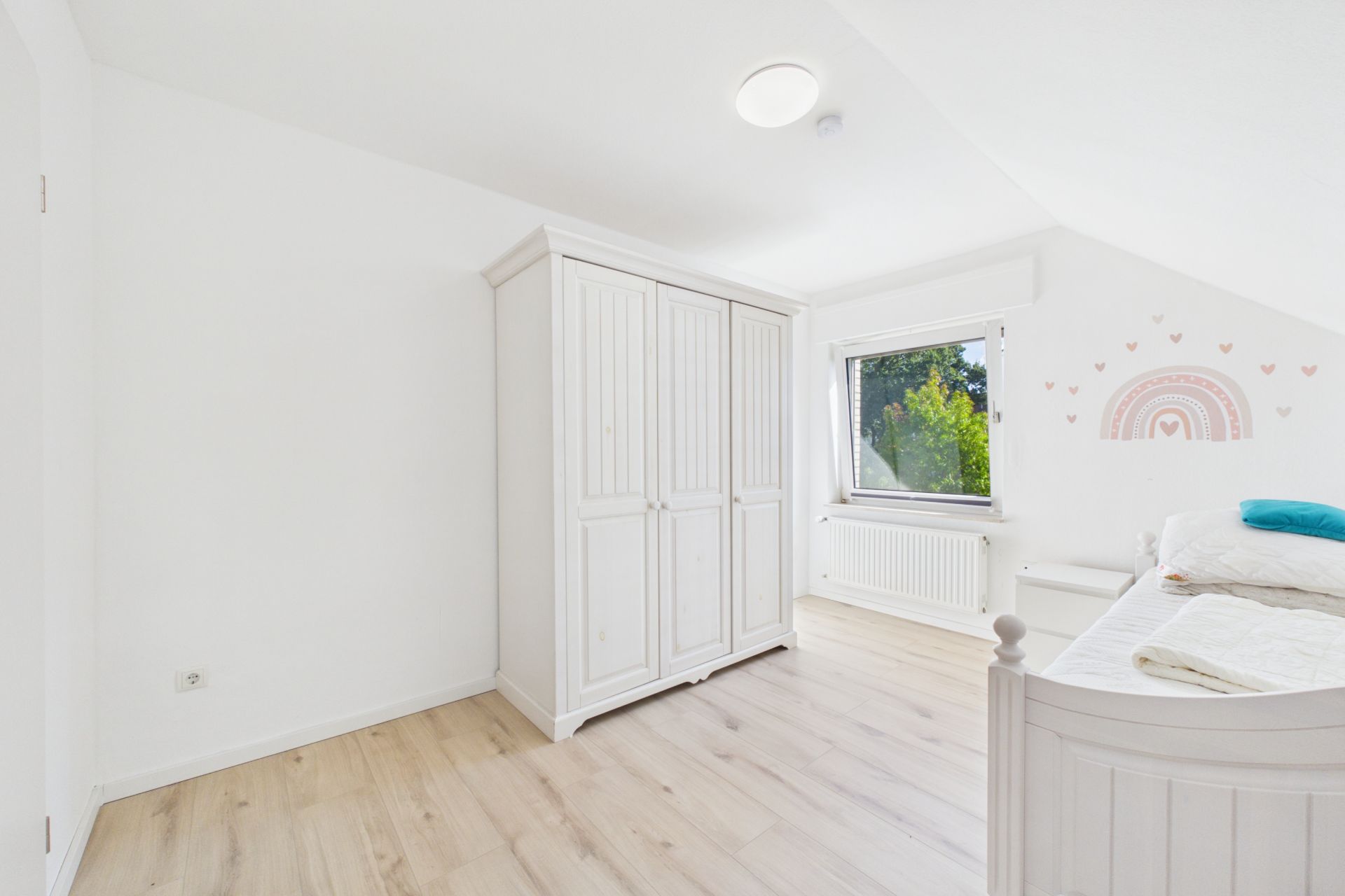 Helles Schlafzimmer mit Holzboden, weißem Schrank, Bett und Fenster mit Blick ins Grüne.