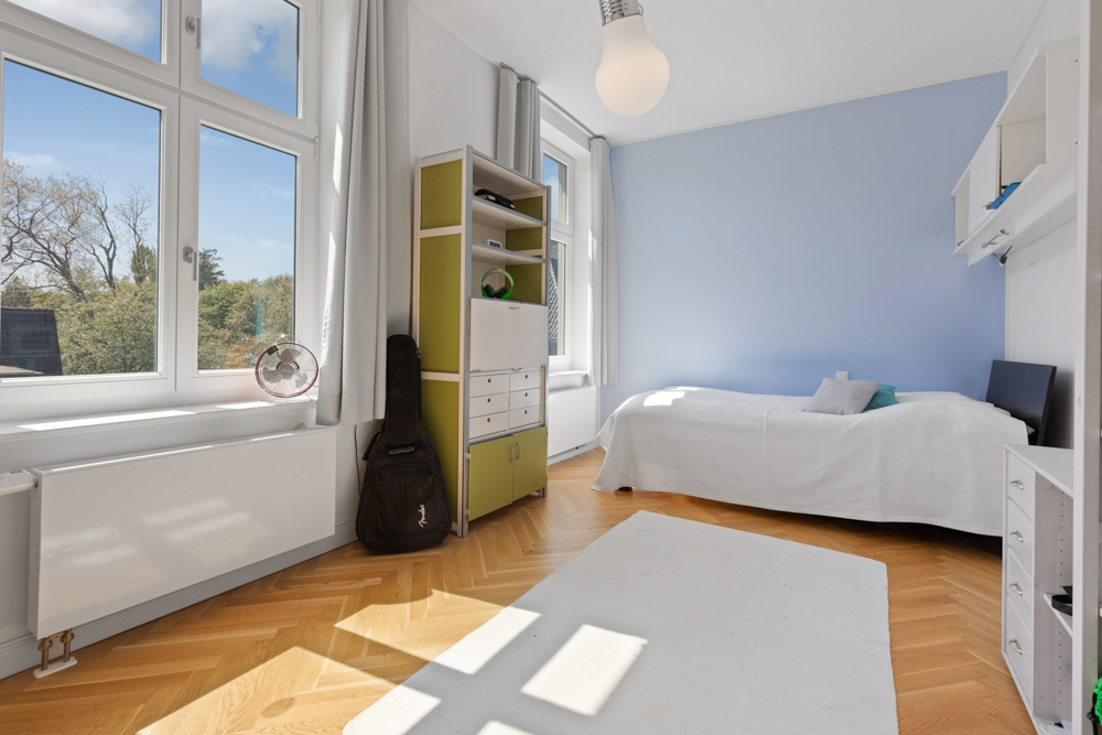 Helles Schlafzimmer mit Einzelbett, großem Fenster, Holzboden, Regal und Gitarre in der Ecke.
