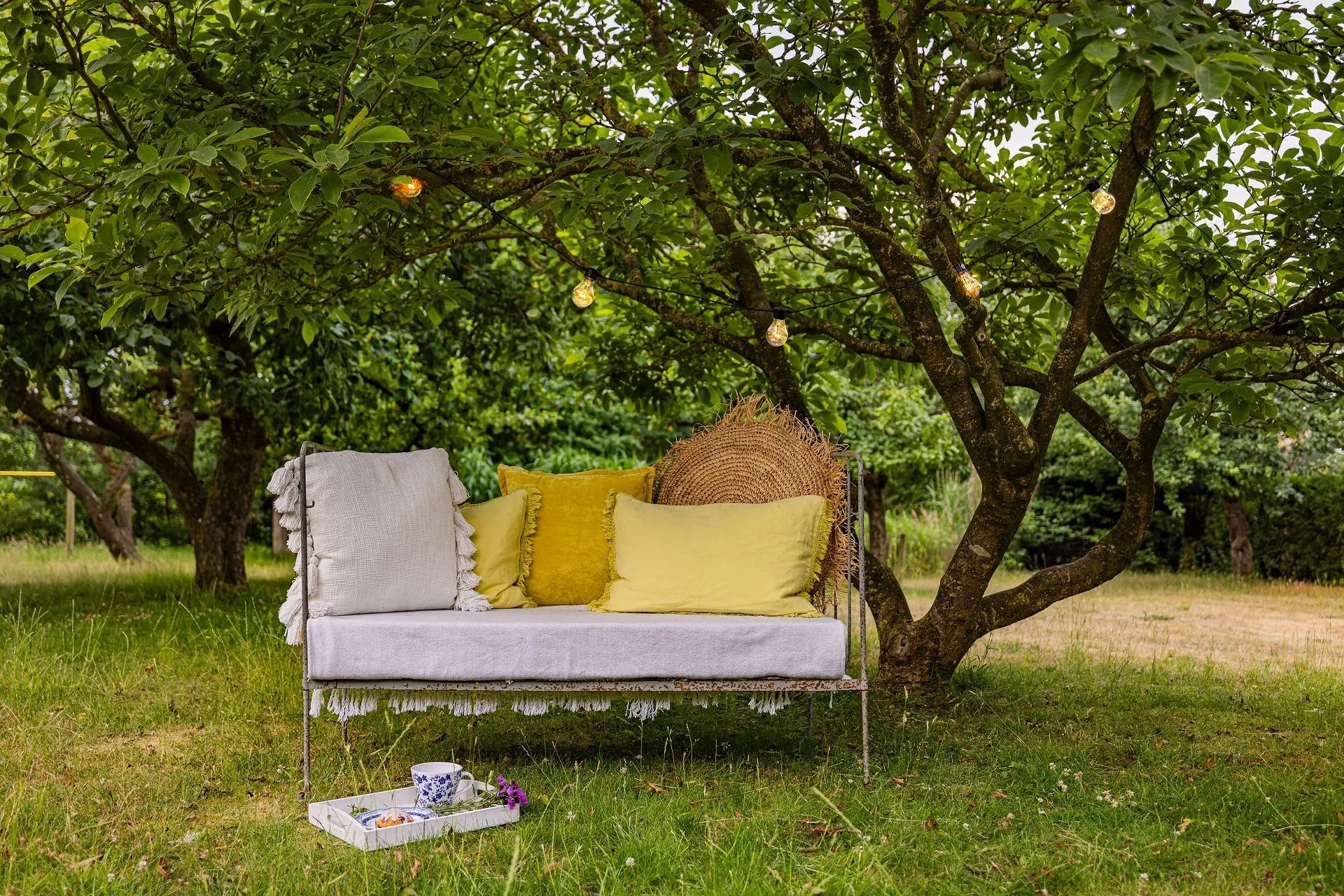 Ein Gartensofa mit Kissen steht unter einem Baum mit Lichterkette, davor ein Tablett mit Tasse.
