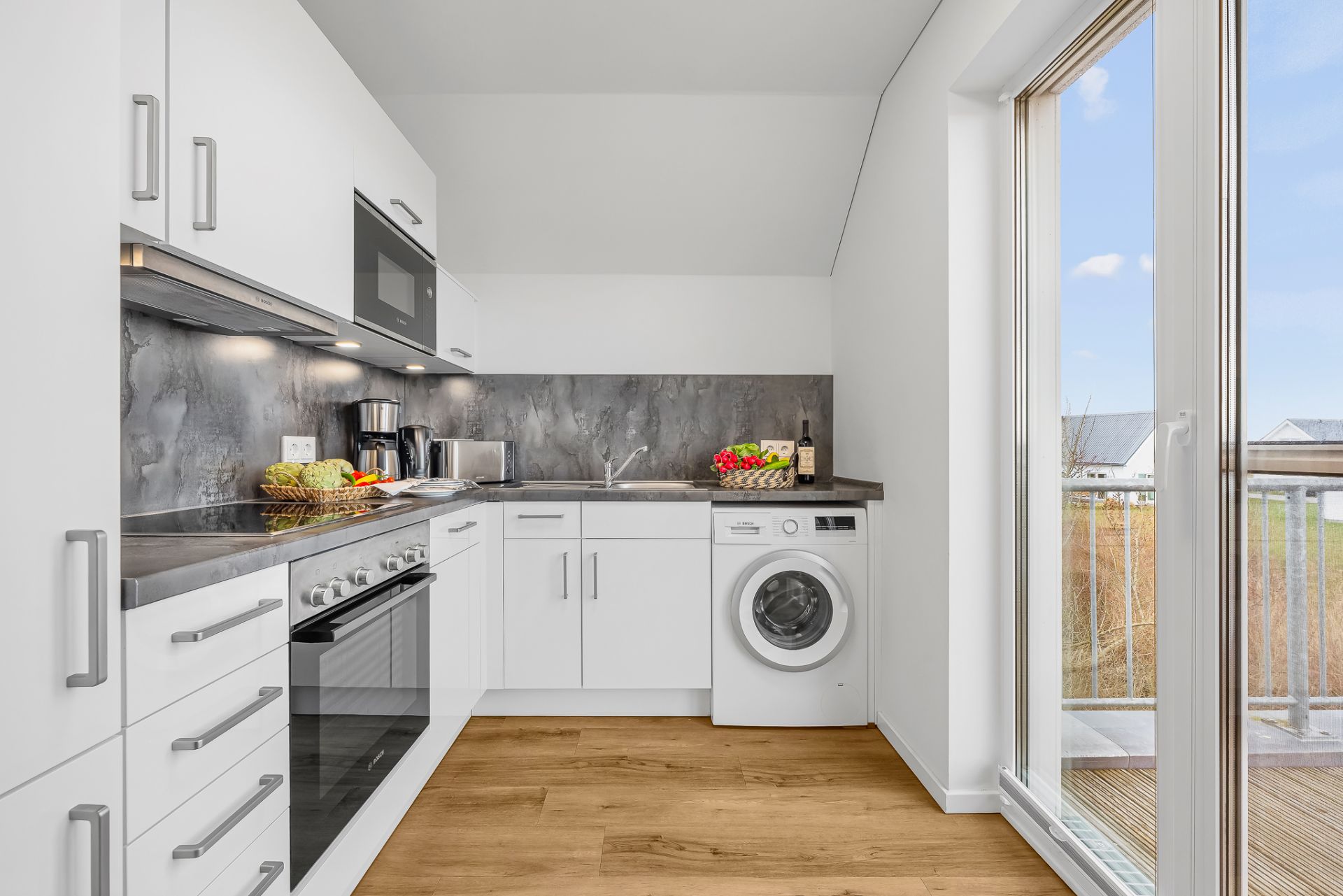 Moderne weiße Küche mit Waschmaschine, Backofen und großem Fenster zum Balkon.