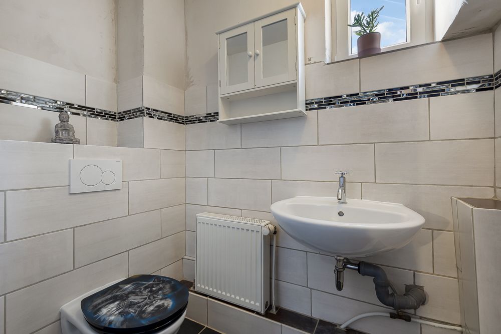 Kleines Bad mit Waschbecken, WC, Heizkörper, Wandregal und Fenster mit Pflanze.