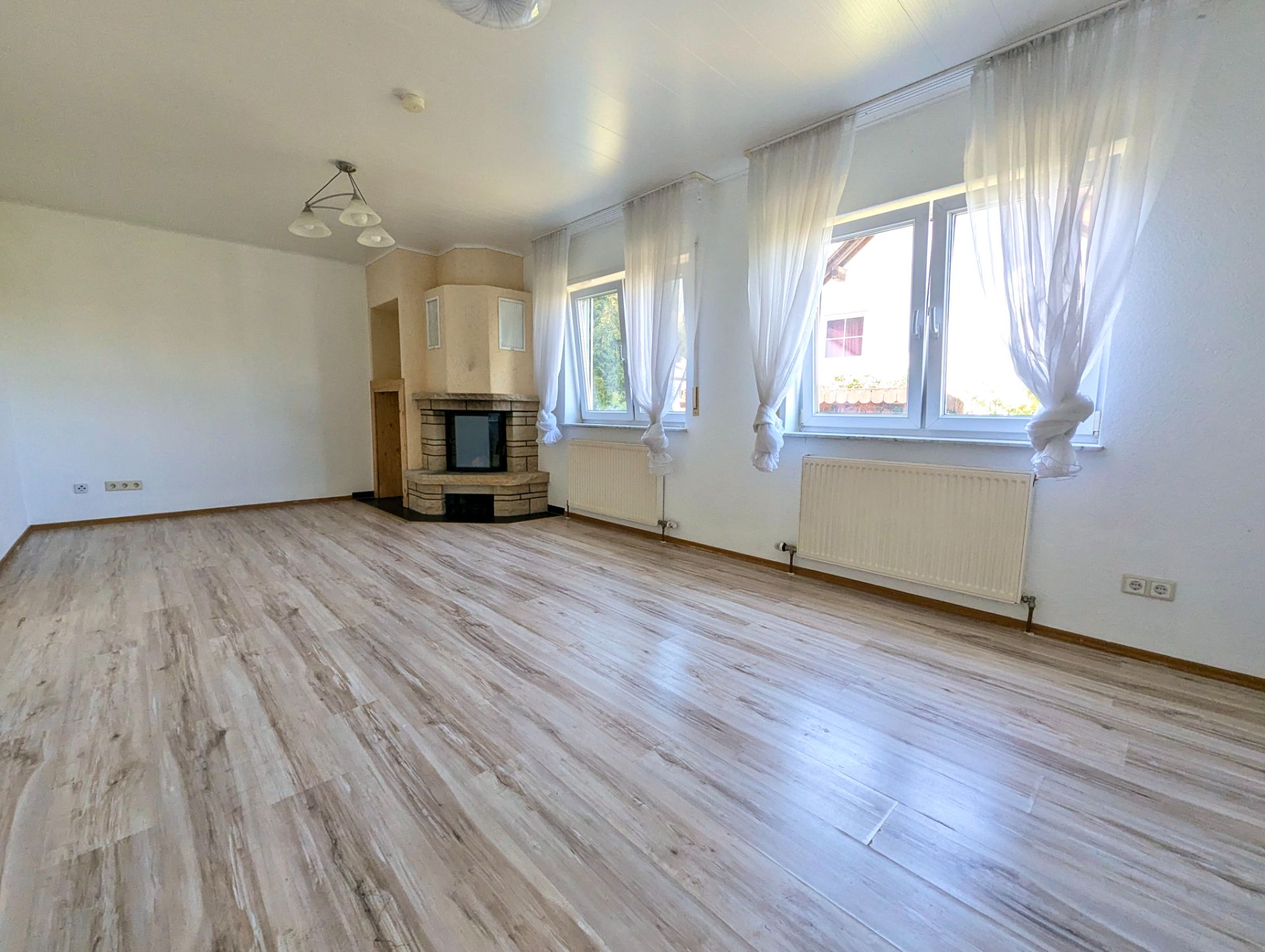 Helles Wohnzimmer mit großen Fenstern, Vorhängen, Kamin und Holzfußboden.