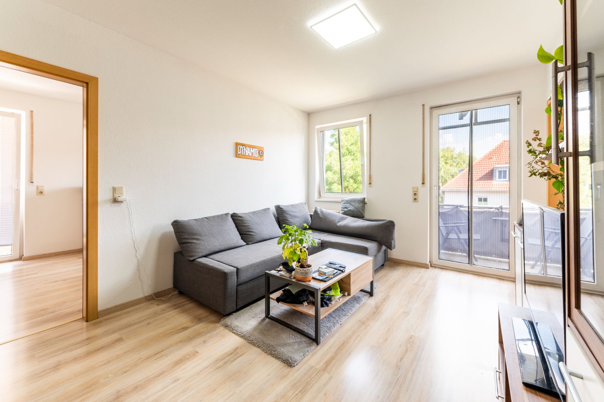 Helles Wohnzimmer mit grauem Ecksofa, Couchtisch, und großem Fenster mit Balkonzugang.