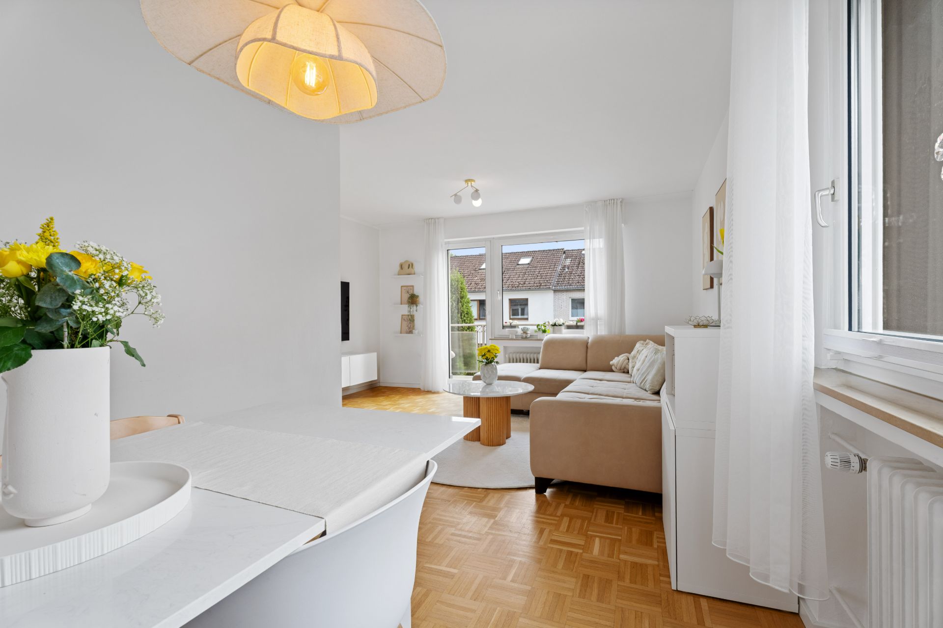 Helles Wohnzimmer mit Ecksofa, Esstisch, Blumen und großen Fenstern mit Ausblick.