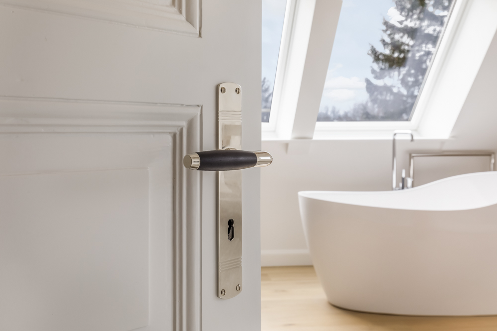 Offene weiße Tür mit silberfarbener Klinke, dahinter eine Badewanne und große Fenster.
