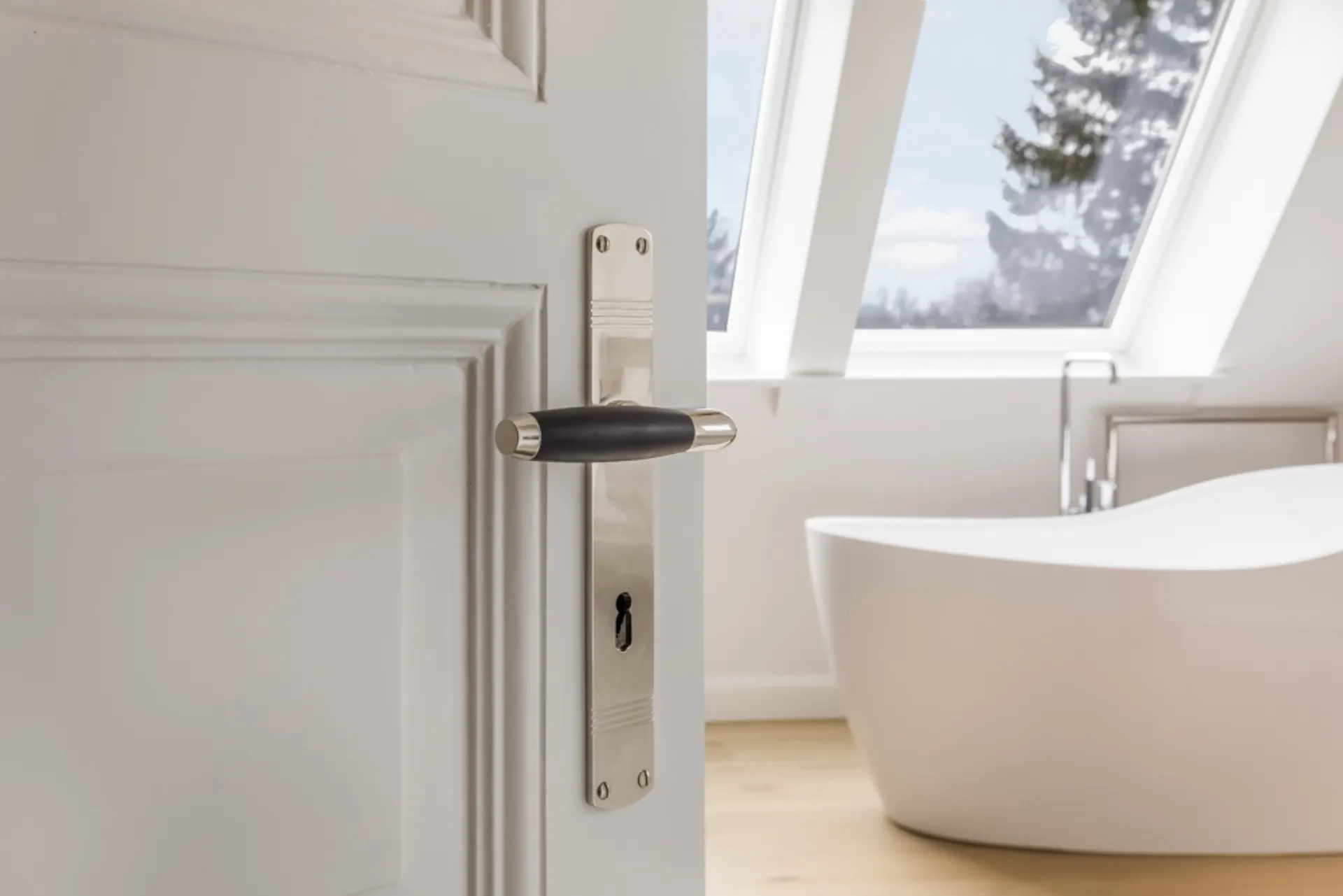 Offene weiße Tür mit silberfarbener Klinke, dahinter eine Badewanne und große Fenster.