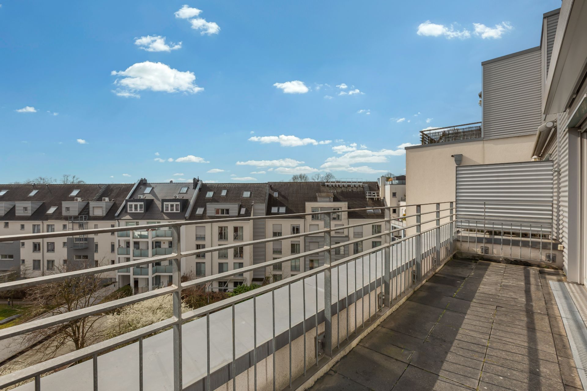 Eine große Terrasse mit Blick auf Wohngebäude und blauen Himmel mit wenigen Wolken.