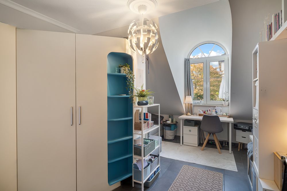 Helles Zimmer mit Schreibtisch am Fenster, Schrank, Regal und moderner Lampe.