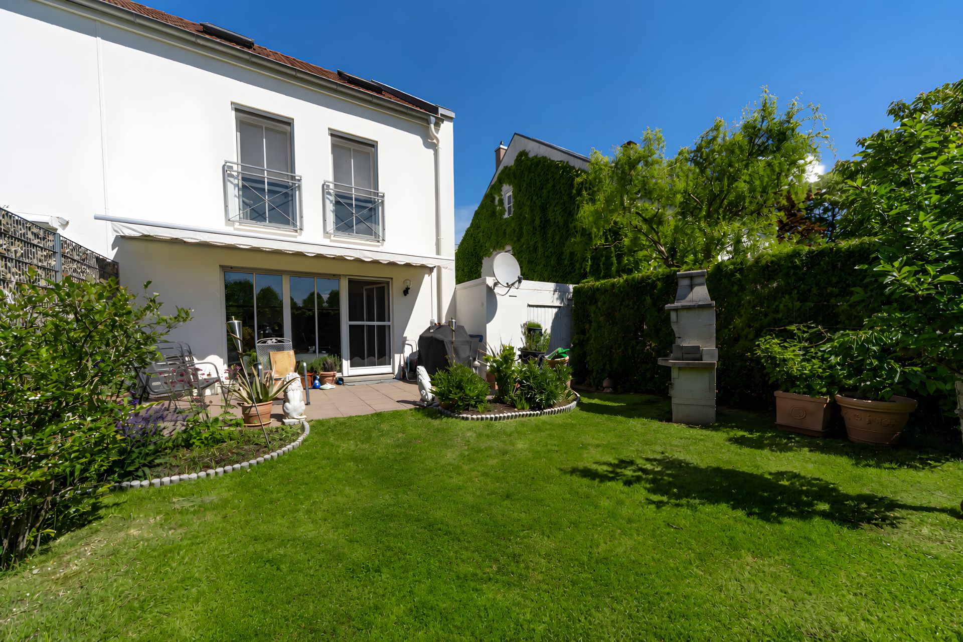 Gepflegter Garten mit Rasen, Terrasse, Pflanzen und Grill vor einem weißen Wohnhaus.