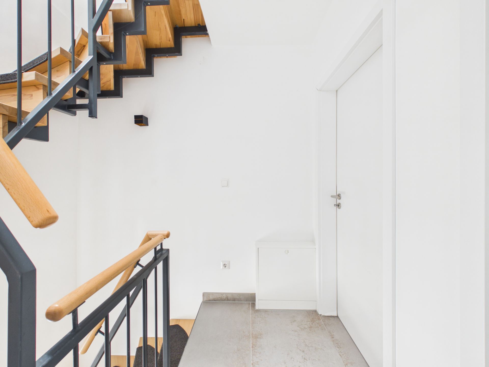 Heller Flur mit Holztreppe und weißer Tür, Geländer mit Holz- und Metallelementen.