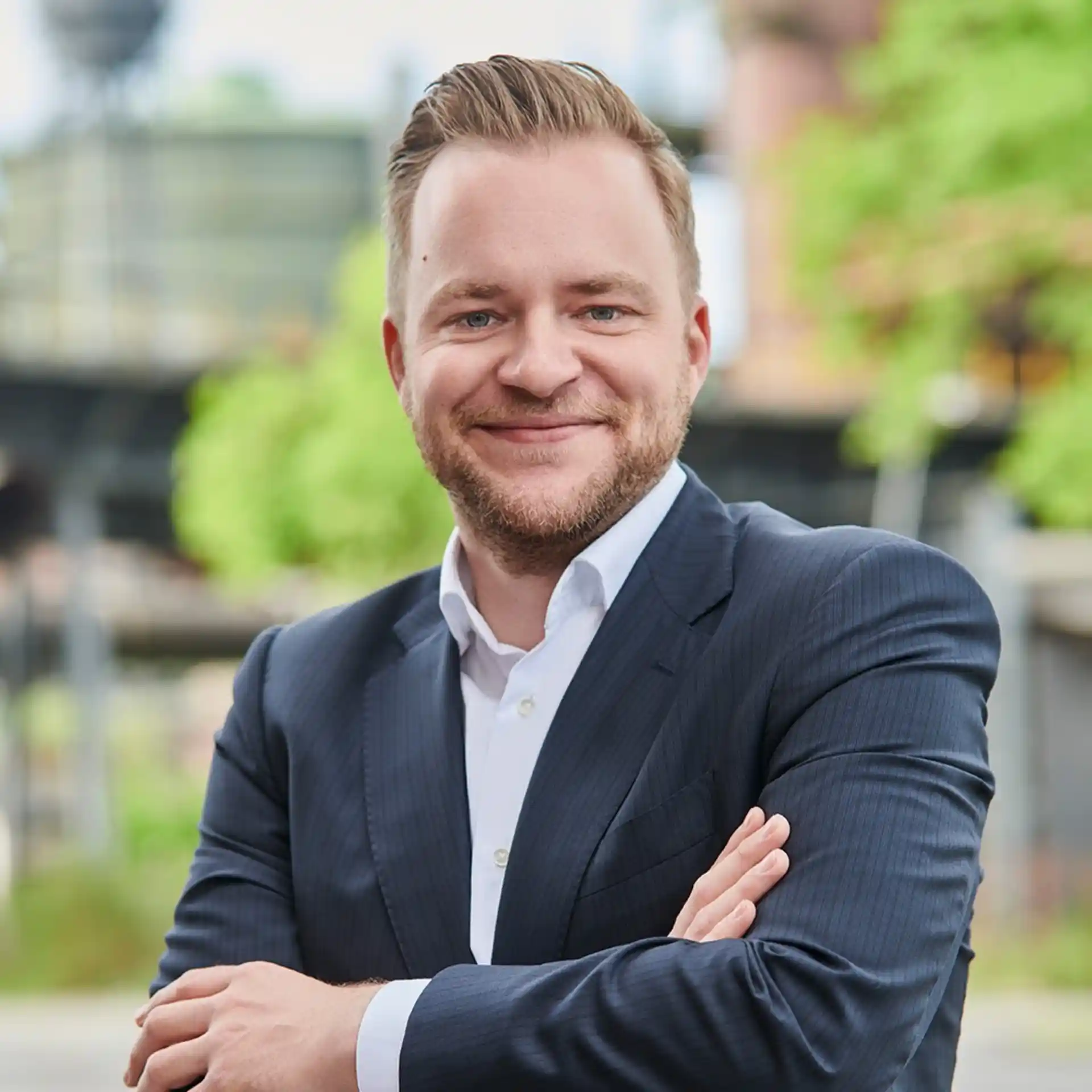 Ein Mann im Anzug steht draußen vor unscharfem, grünem Hintergrund und lächelt.
