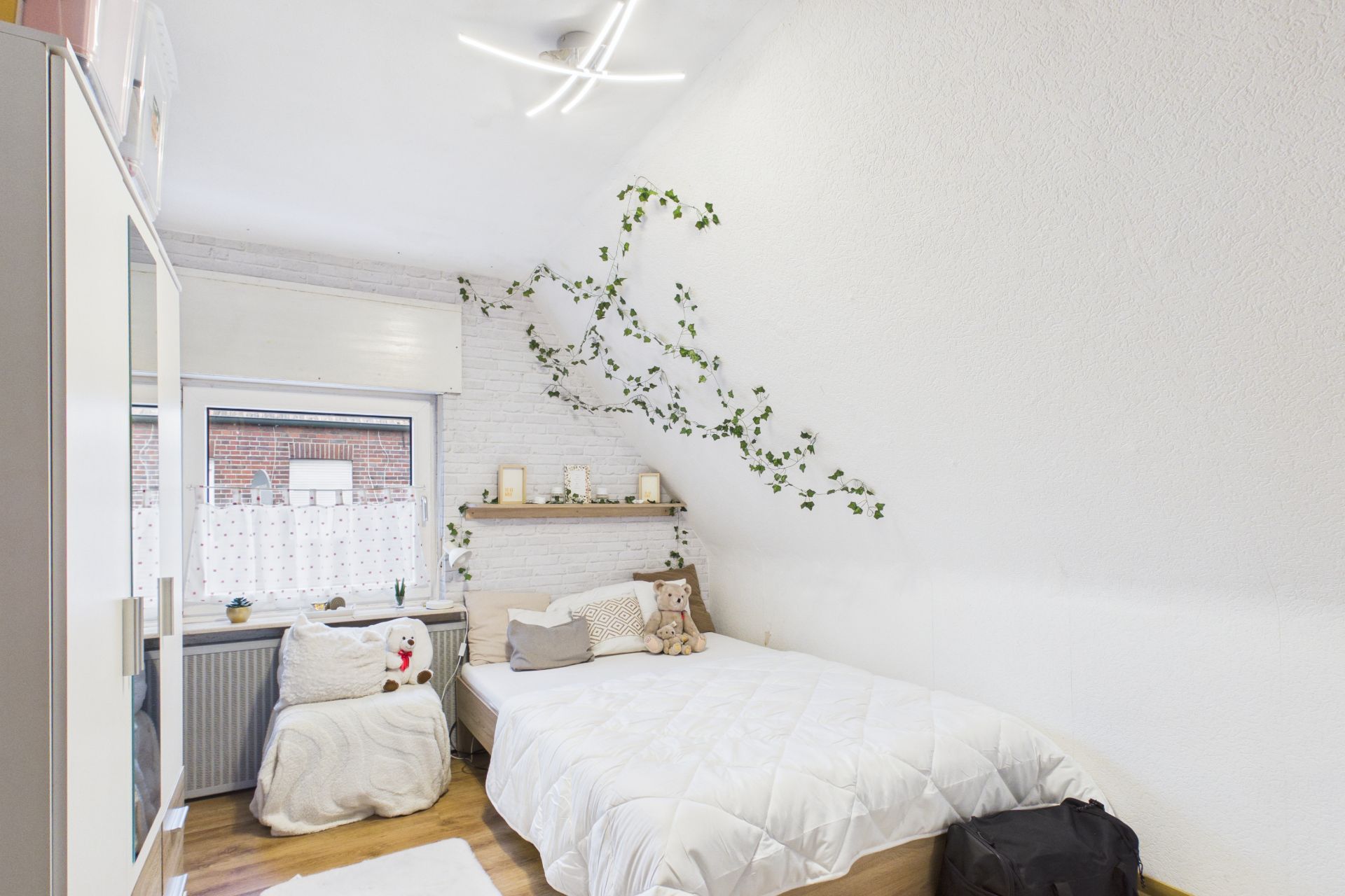 Helles Schlafzimmer mit Bett, Sessel, Fenster und dekorativen Pflanzen an der weißen Wand.