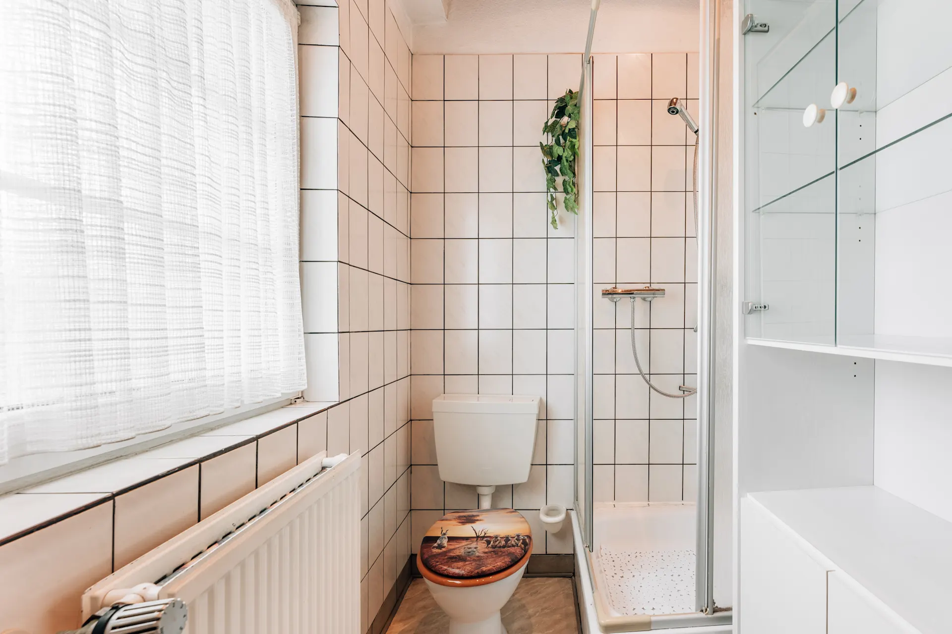 Kleines Badezimmer mit Toilette, Dusche, Heizkörper und Regal, gefliest in Weiß.