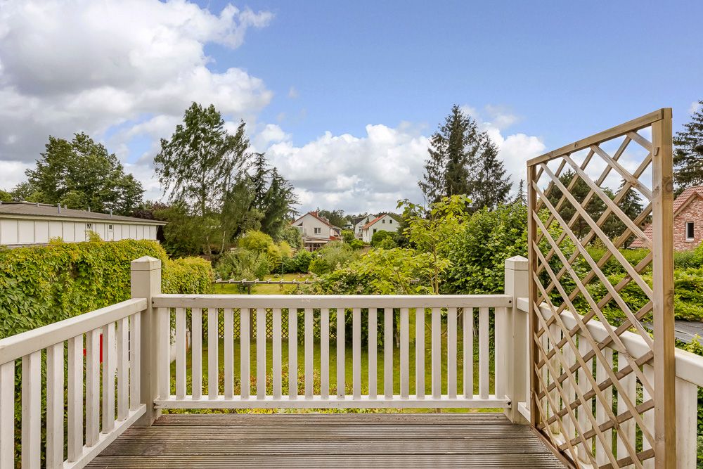 Holzbalkon mit weißem Geländer und seitlichem Rankgitter, Blick auf grünen Garten und Häuser.