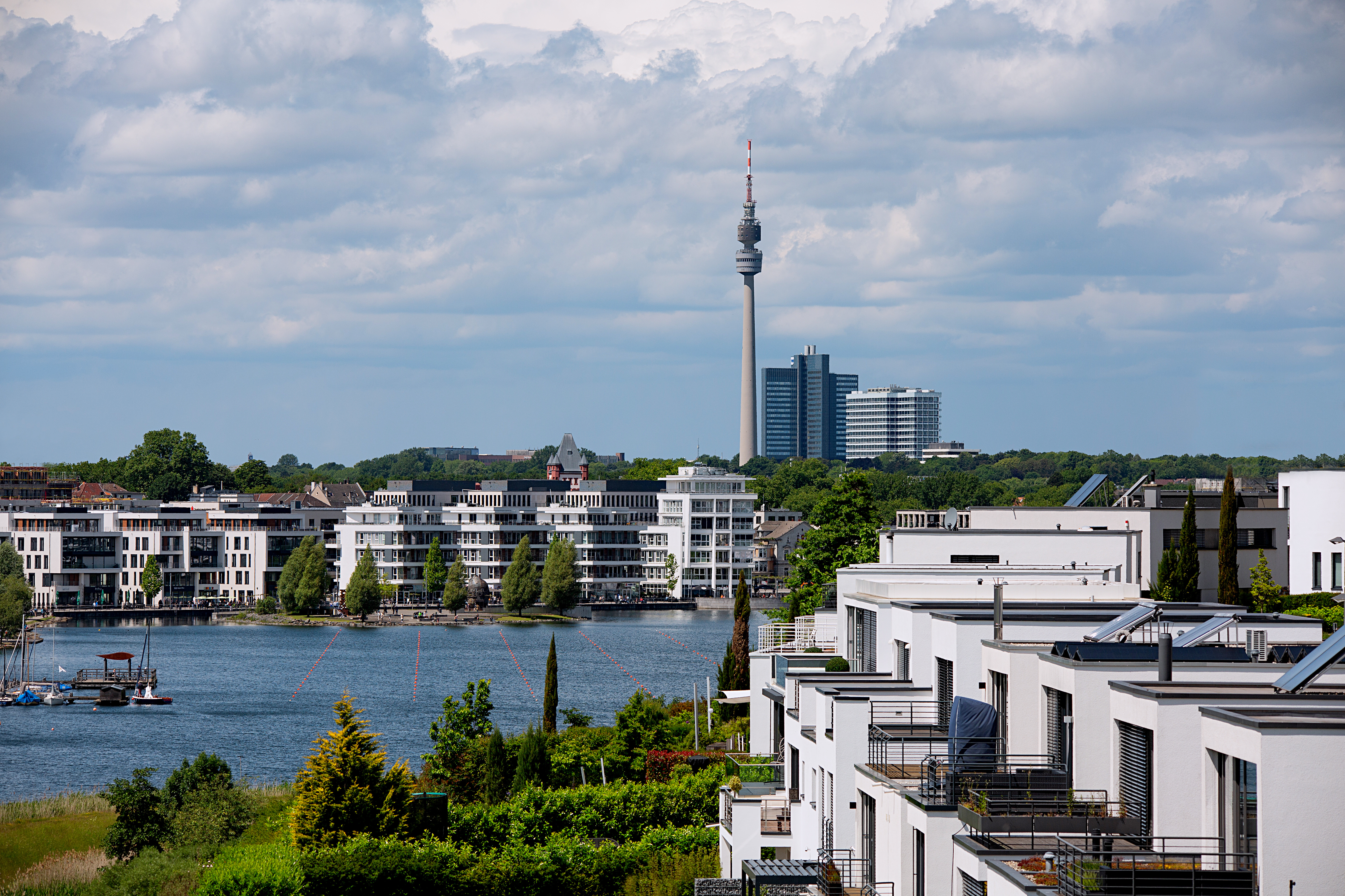 Blick auf moderne Wohnhäuser am Wasser mit einem Fernsehturm und Hochhäusern im Hintergrund.