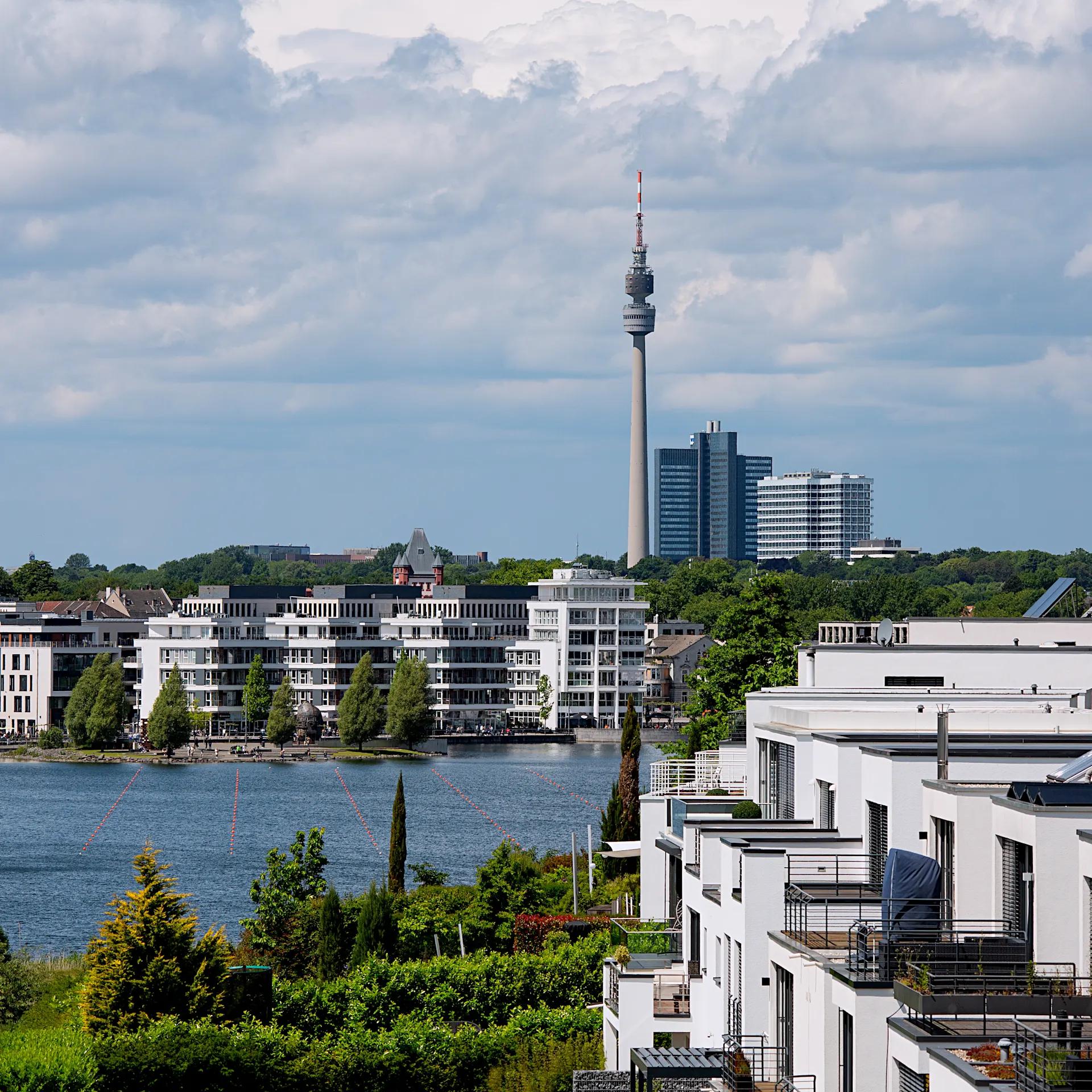 Blick auf moderne Wohnhäuser am Wasser mit einem Fernsehturm und Hochhäusern im Hintergrund.