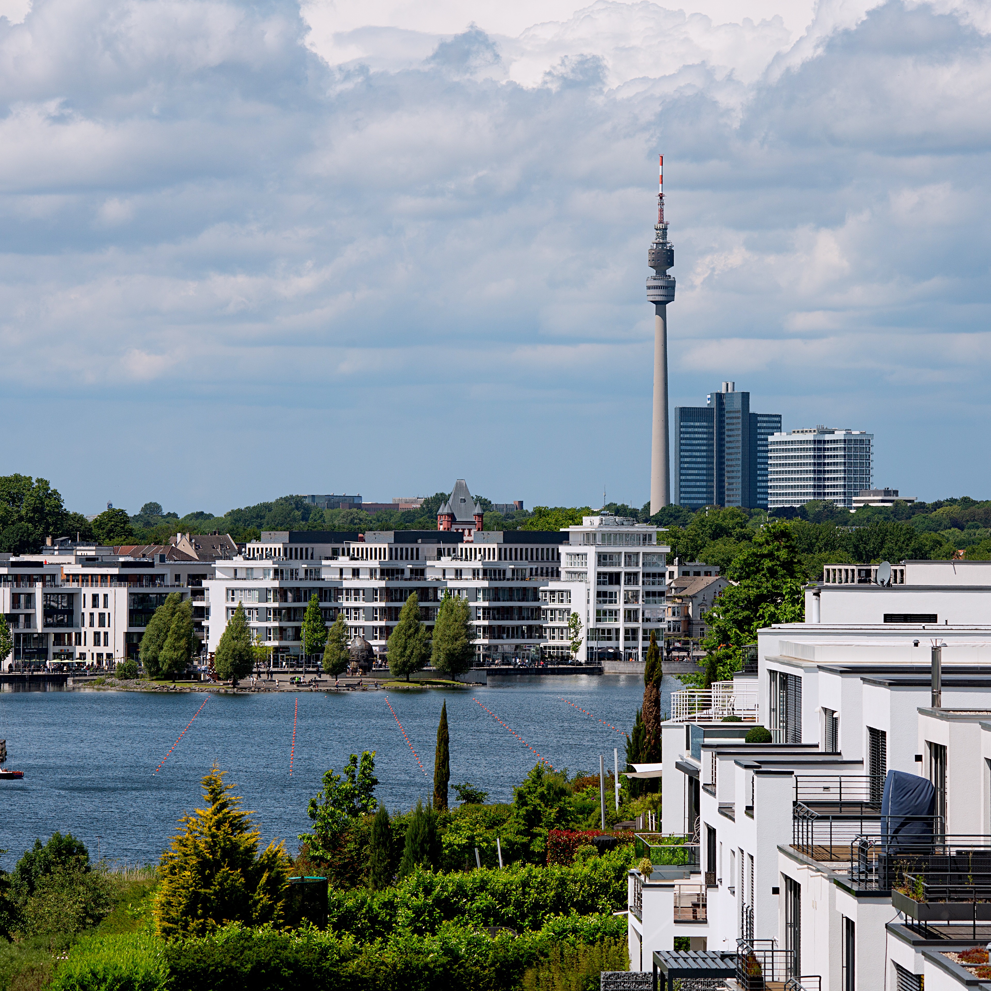 Blick auf moderne Wohnhäuser am Wasser mit einem Fernsehturm und Hochhäusern im Hintergrund.