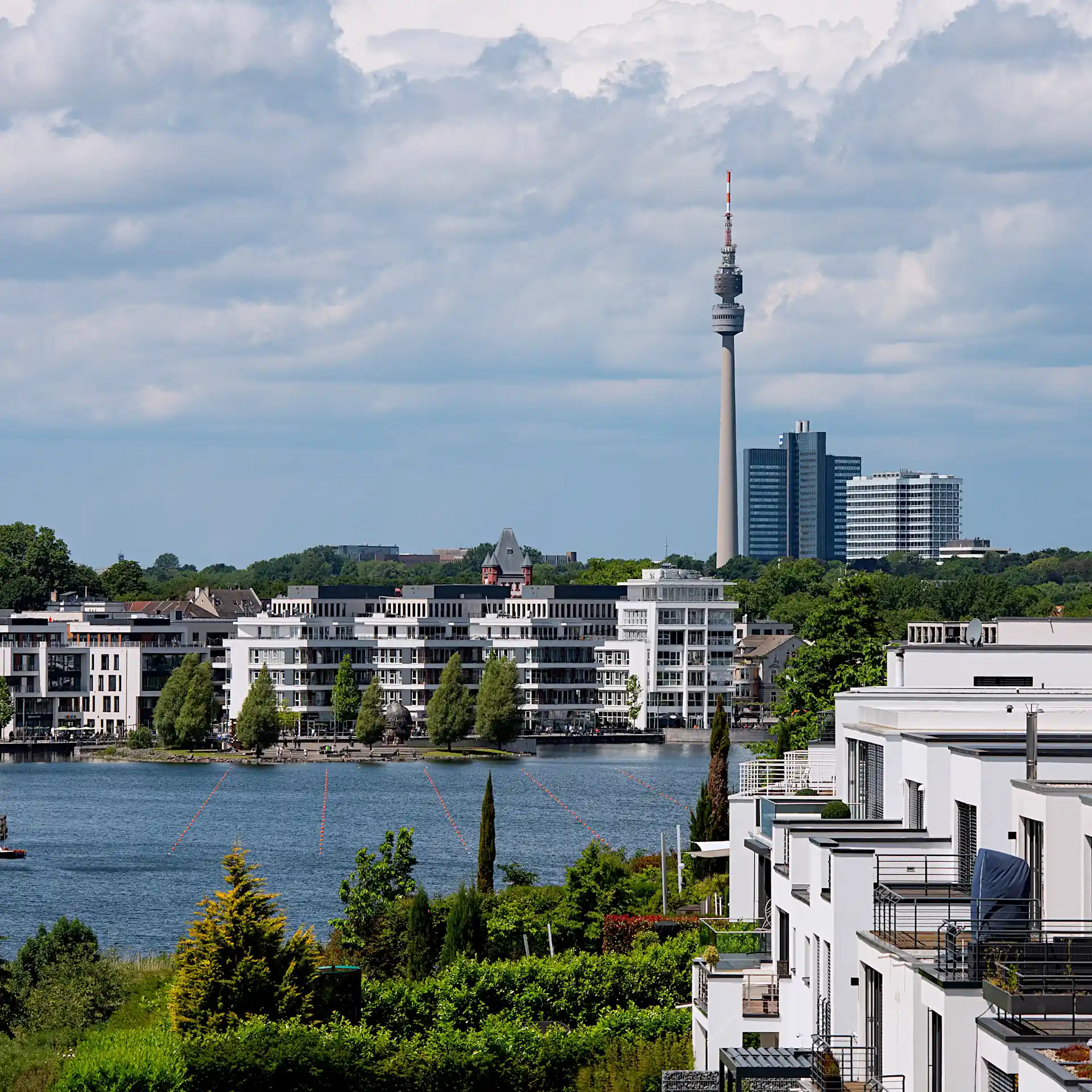 Blick auf moderne Wohnhäuser am Wasser mit einem Fernsehturm und Hochhäusern im Hintergrund.