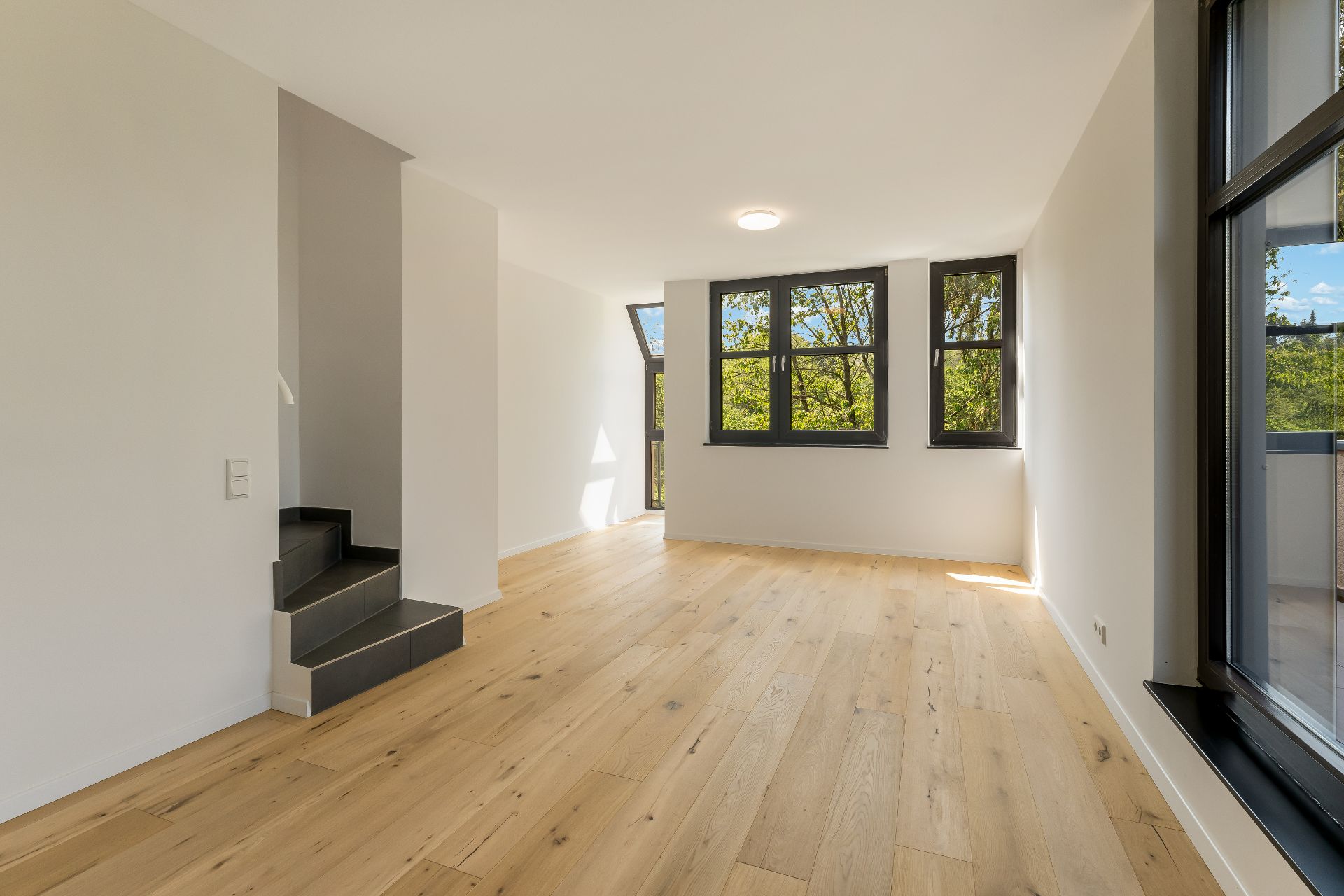 Heller Raum mit Holzfußboden, großen Fenstern und einer schwarzen Treppe.