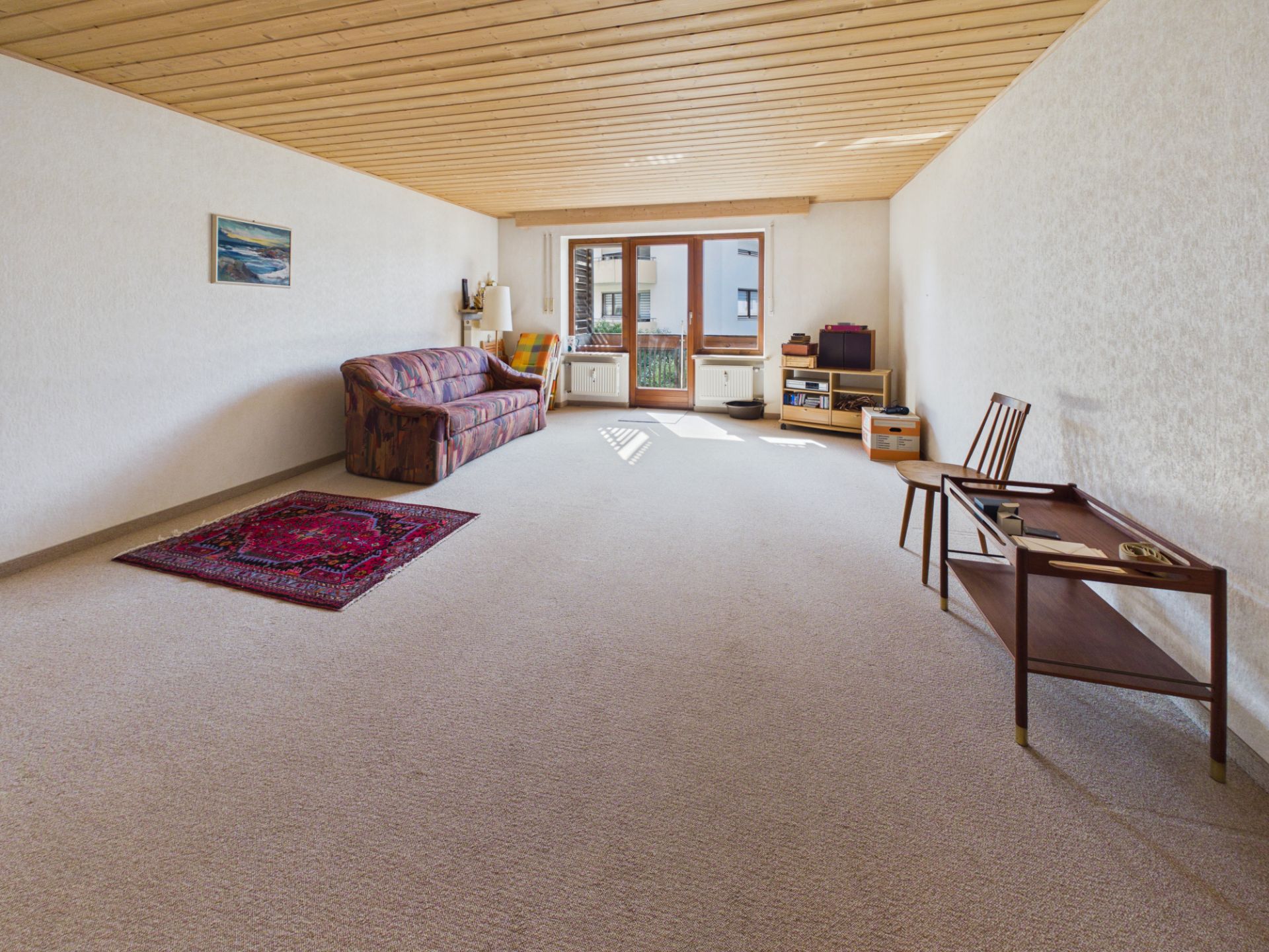Helles Wohnzimmer mit Sofa, Teppich, Stuhl und großem Fenster mit Zugang zum Balkon.