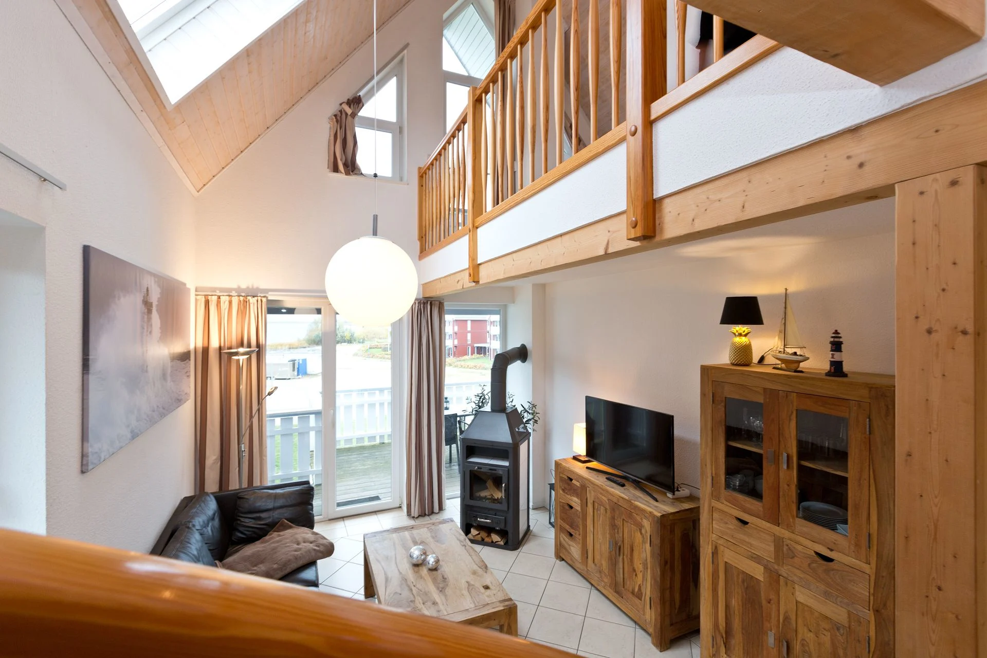 Wohnzimmer mit Holzdecke, Sofa, Fernseher, Kaminofen und Holzmöbeln, Galerie im Obergeschoss.