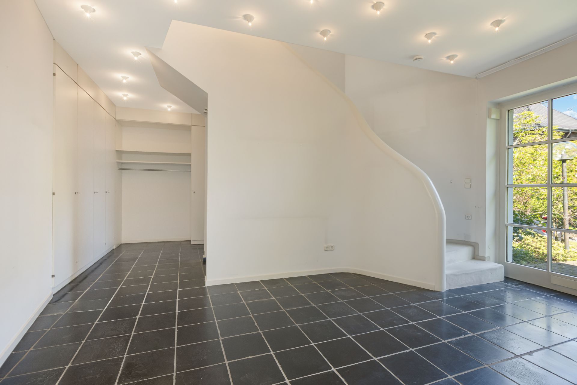 Heller Raum mit schwarzen Fliesen, weiße Einbauschränke und geschwungene Treppe am Fenster.