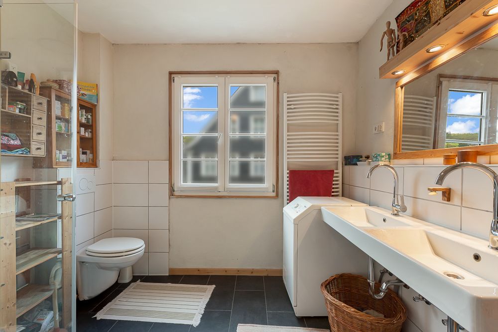 Helles Badezimmer mit Doppelwaschbecken, Waschmaschine, WC, Dusche und Fenster.