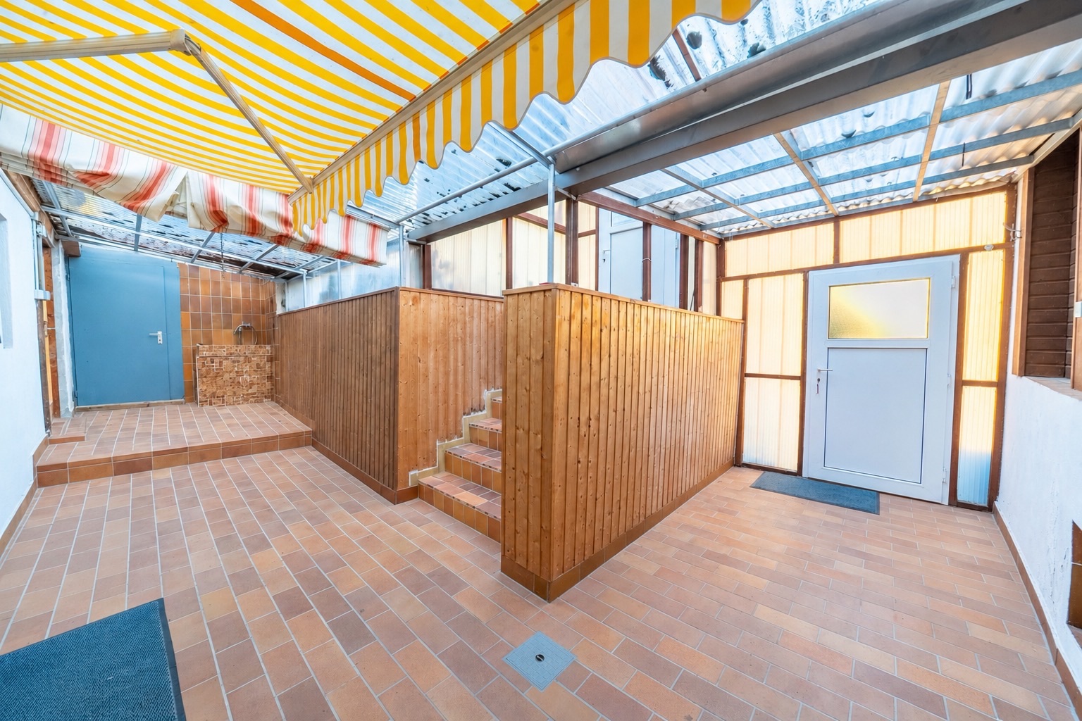 Überdachter Innenhof mit Fliesenboden, Holzverkleidung und gelb-weißer Markise.