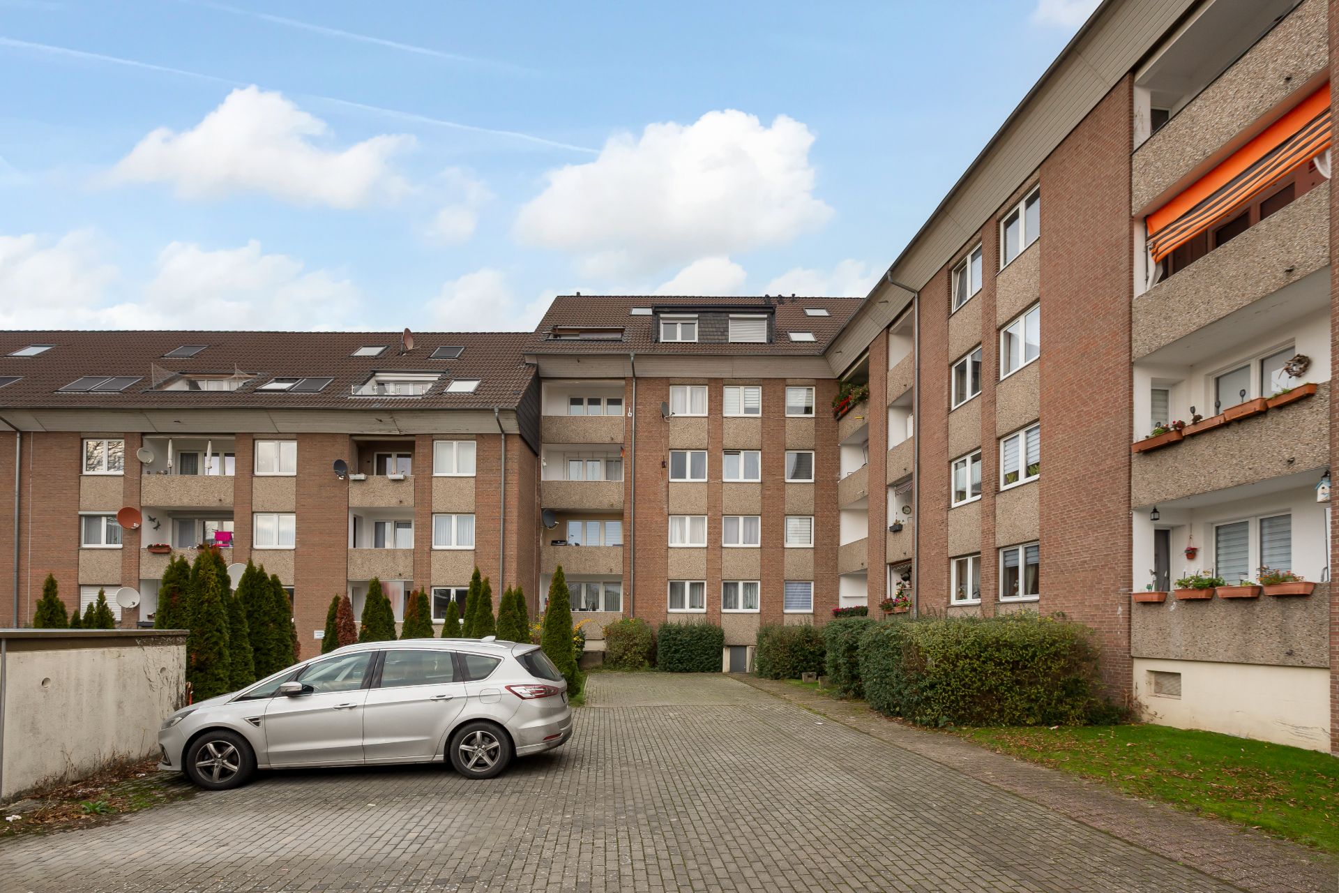 Mehrfamilienhaus mit mehreren Wohnungen, davor ein graues Auto auf gepflastertem Hof.