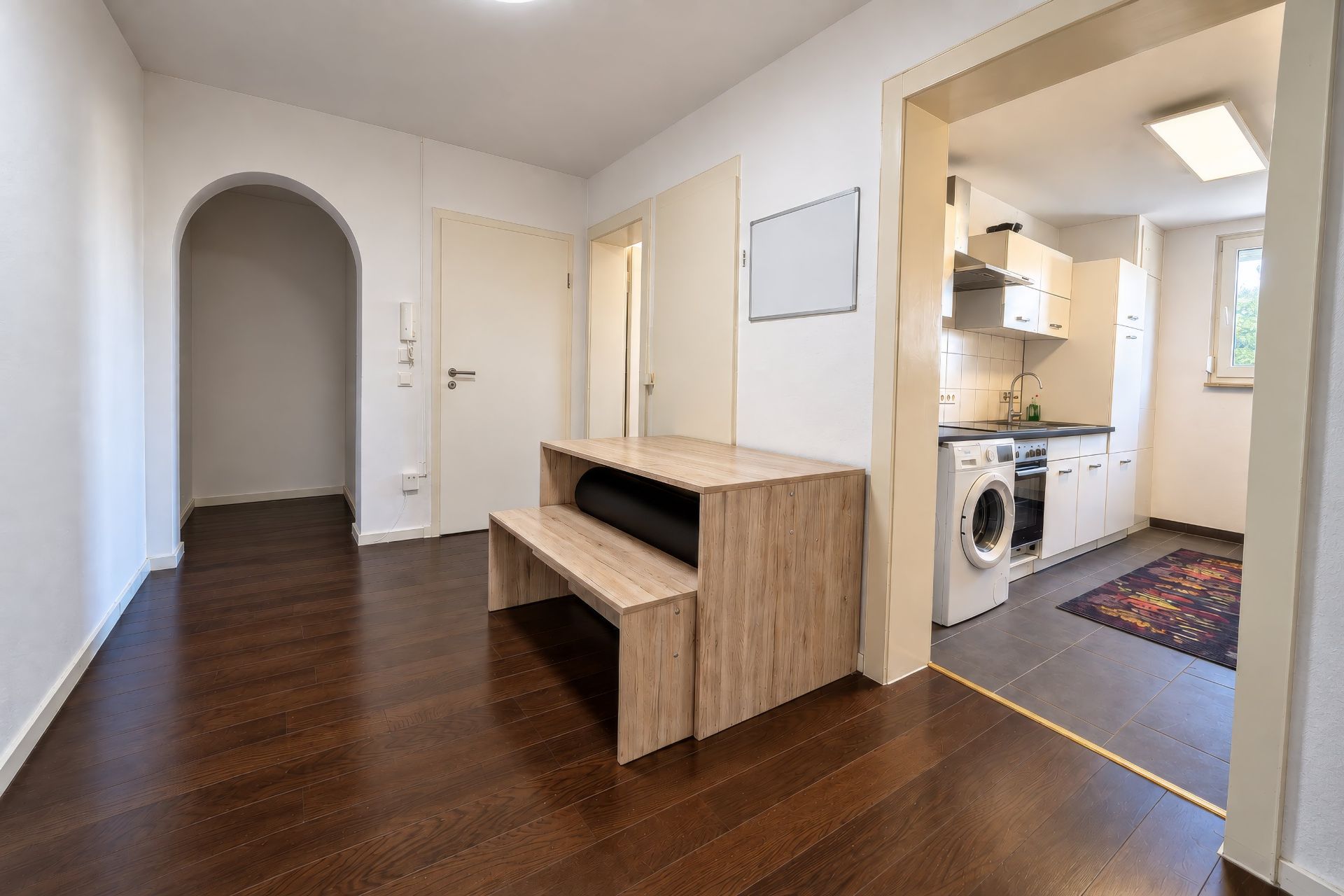 Flur mit dunklem Holzboden, Tisch mit Sitzbank und Blick in eine Küche mit Waschmaschine.