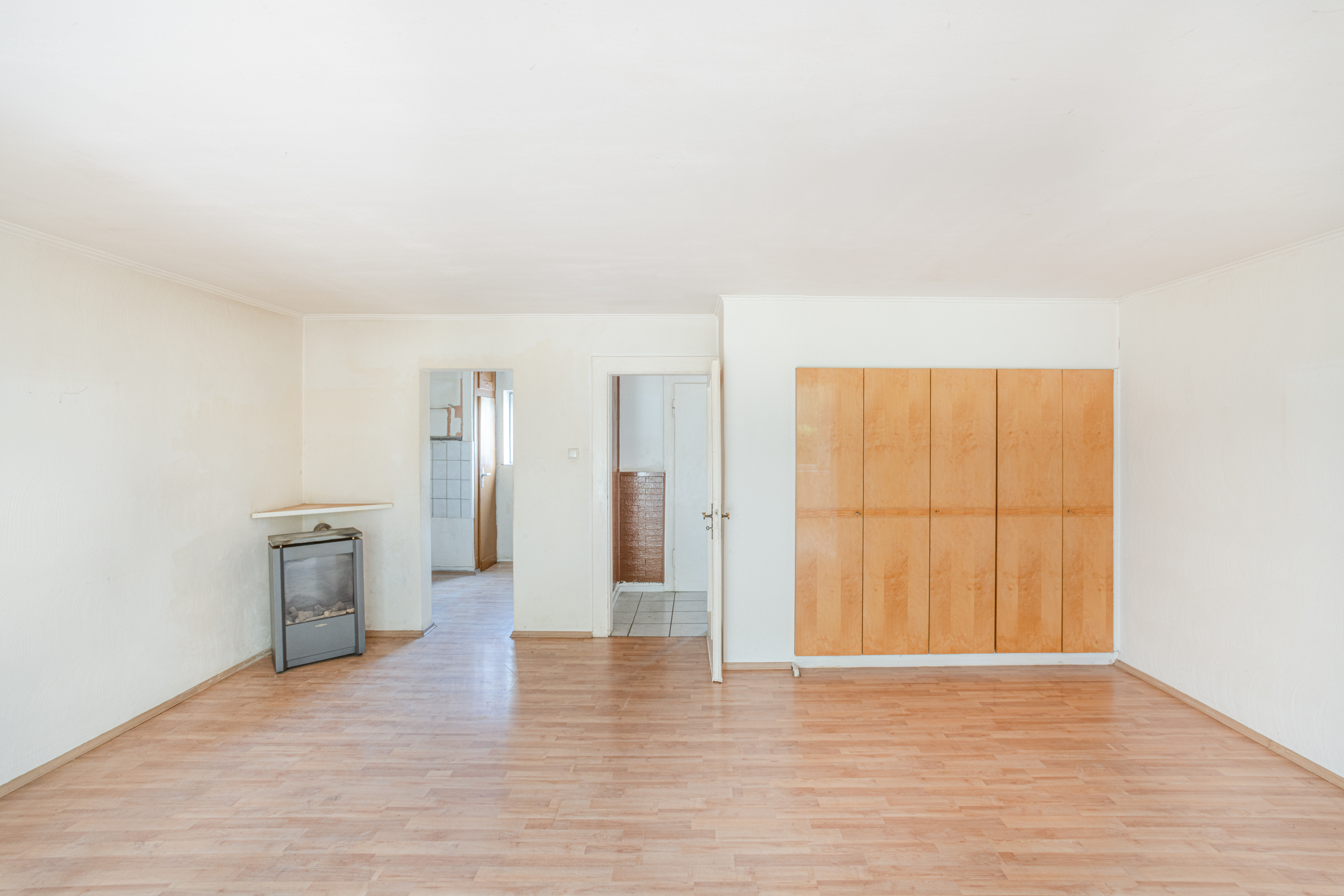 Heller leerer Raum mit Holzschrank, Ofen und Türen zu angrenzenden Zimmern.