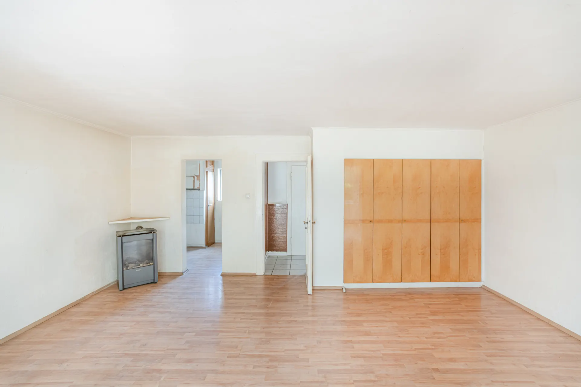 Heller leerer Raum mit Holzschrank, Ofen und Türen zu angrenzenden Zimmern.