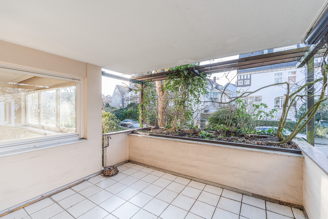Überdachter Balkon mit Fliesenboden, Fenster und Pflanzbeeten mit Kletterpflanzen.