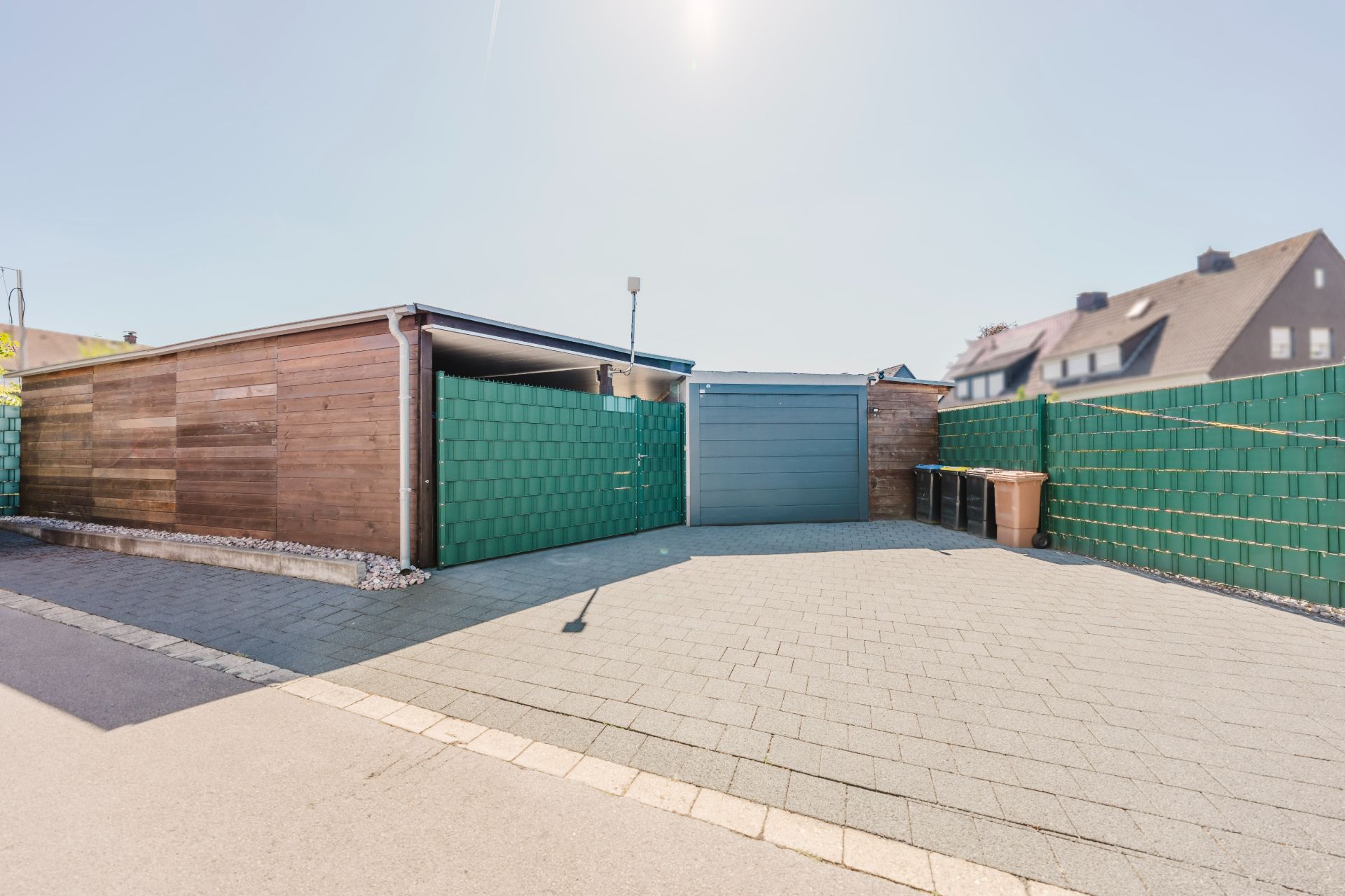 Gepflasterter Hof mit Garage, Mülltonnen und grüner Zaunanlage bei Wohnhäusern.
