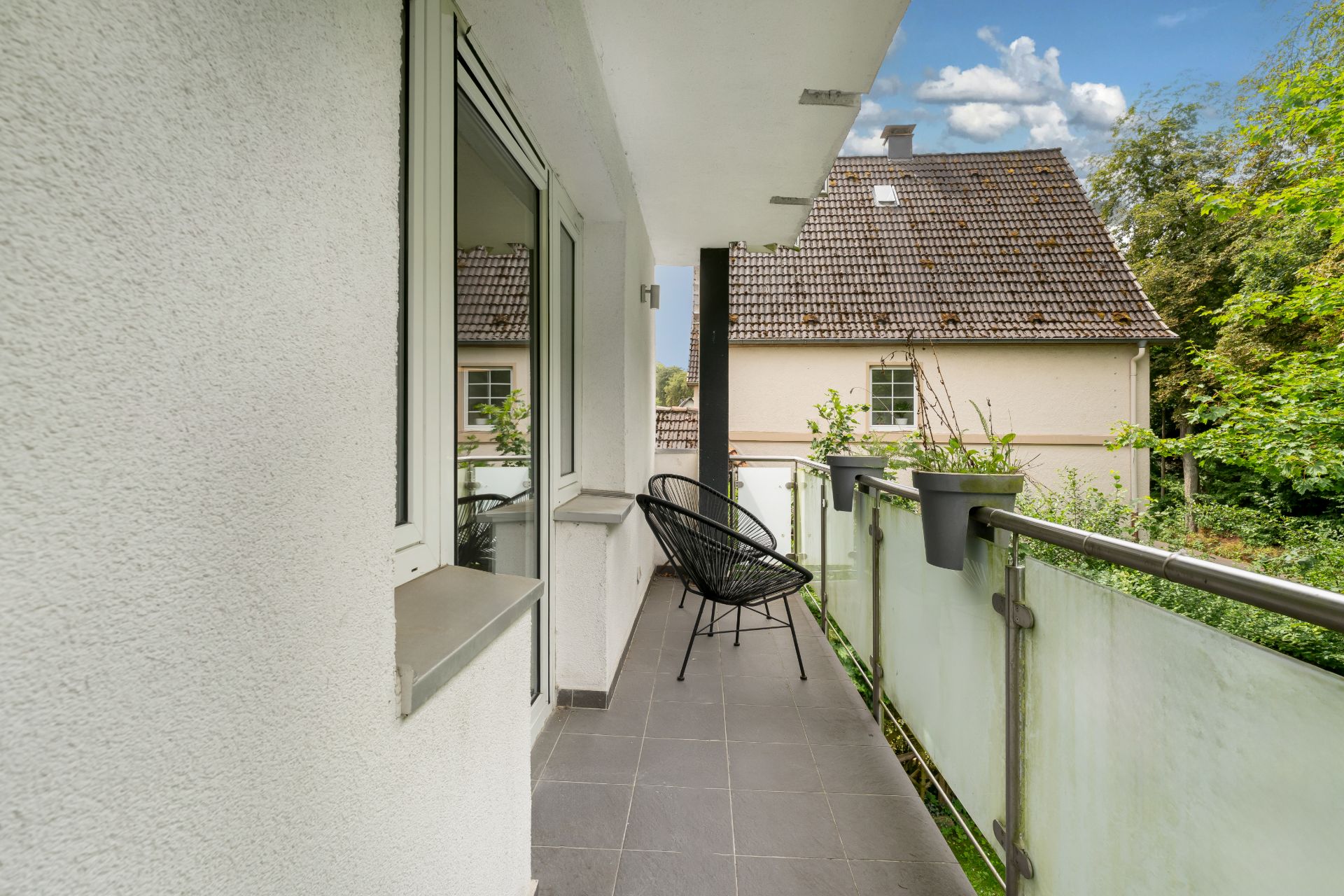 Balkon mit Glasgeländer, einem Stuhl und Blick auf ein Nachbarhaus und Bäume.