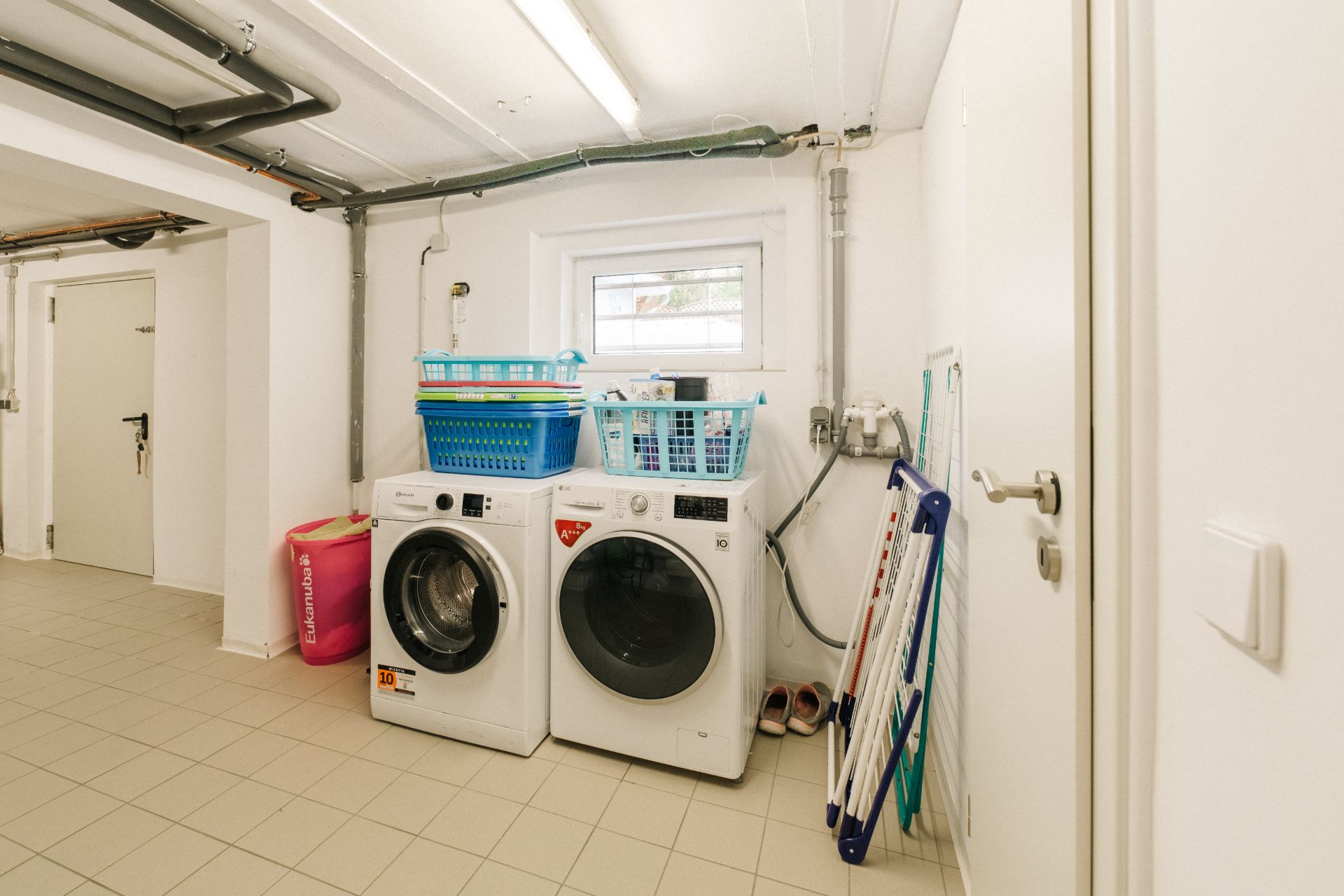 Waschraum mit Waschmaschine, Trockner, Wäschekörben und Wäschetrockner neben einem Fenster.