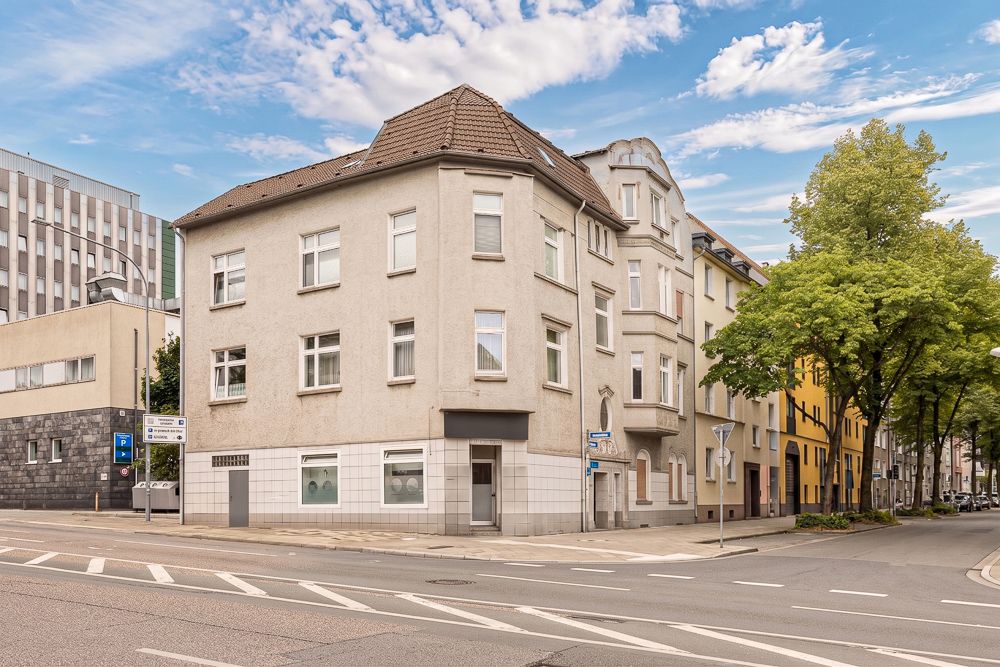 Eckhaus mit mehreren Fenstern und Eingangstür an einer Straßenkreuzung, Bäume am Gehweg.