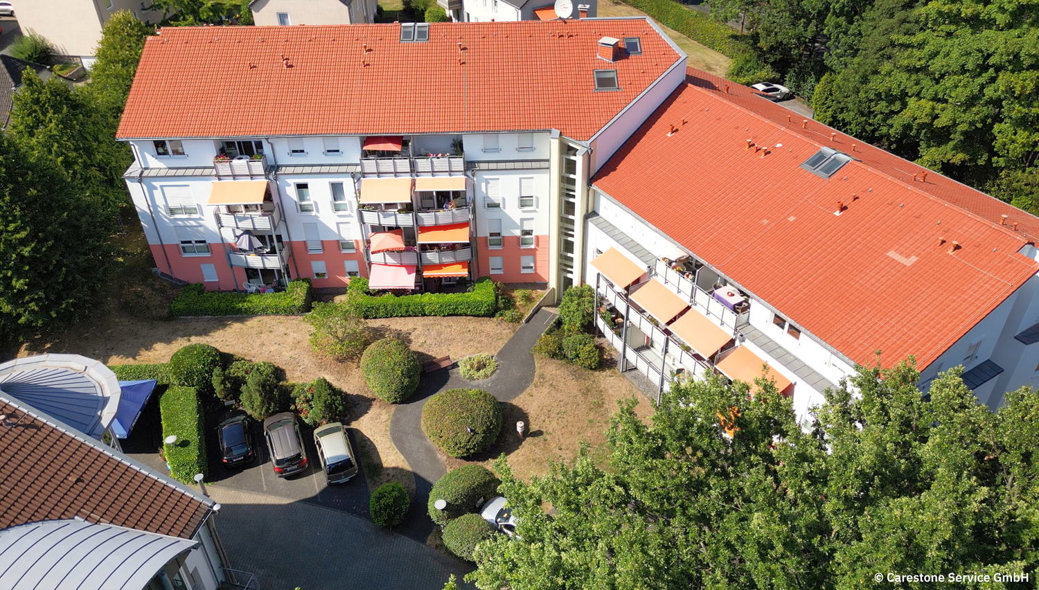 Wohnhaus mit roten Dächern, Balkonen und kleinem Garten mit Gehwegen und parkenden Autos.