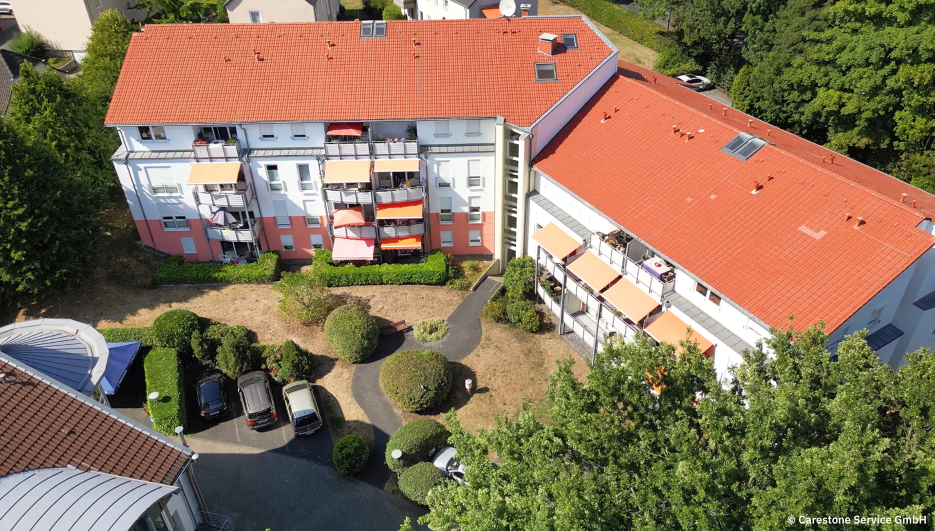 Wohnhaus mit roten Dächern, Balkonen und kleinem Garten mit Gehwegen und parkenden Autos.