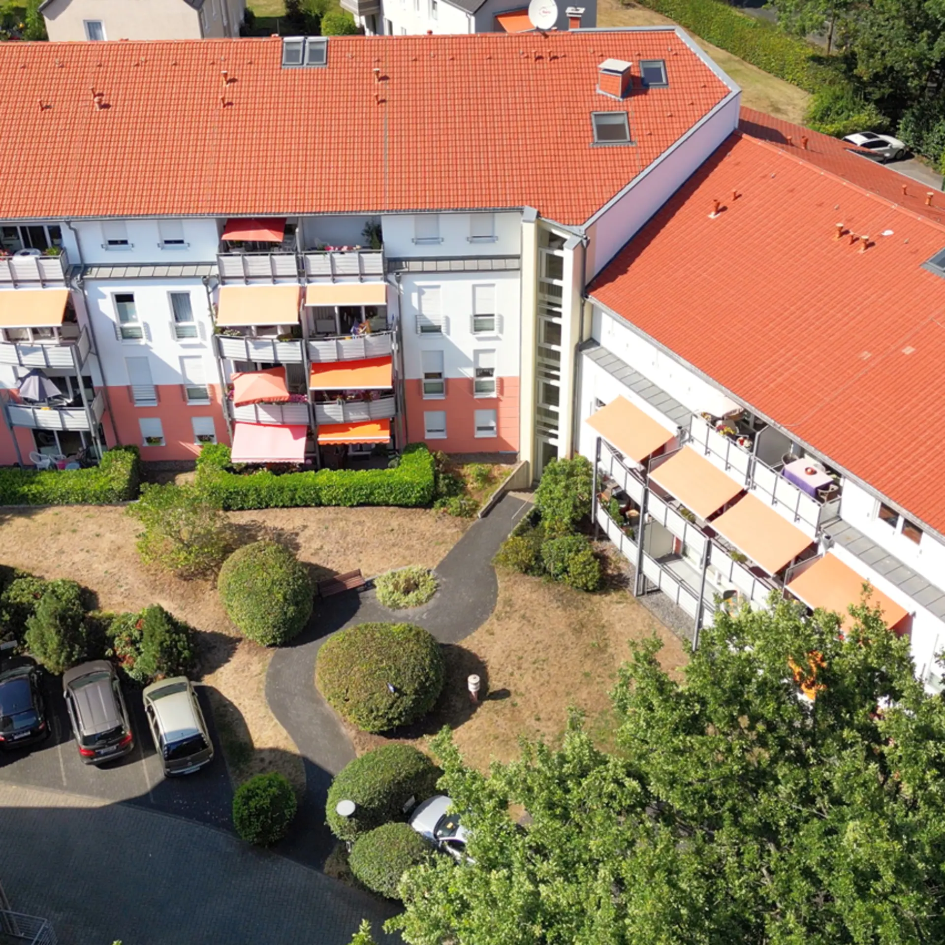 Wohnhaus mit roten Dächern, Balkonen und kleinem Garten mit Gehwegen und parkenden Autos.