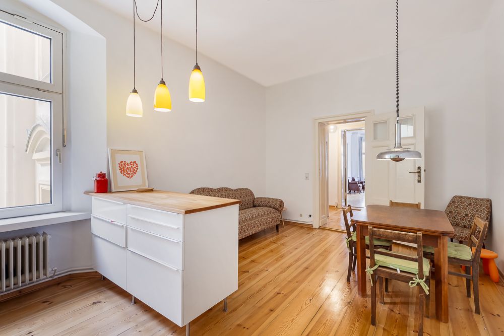 Helles Wohnzimmer mit Holzboden, Sofa, Esstisch und Kücheninsel an einem Fenster.