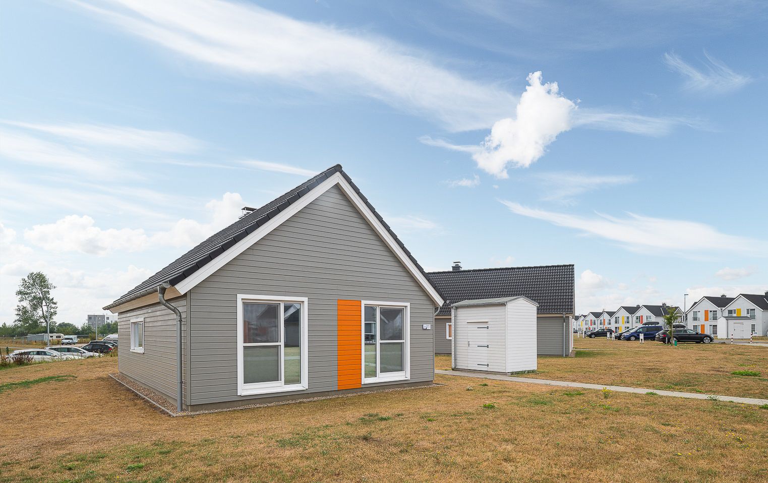 Graues Einfamilienhaus mit Schrägdach, großen Fenstern und orangefarbener Tür auf trockenem Rasen.