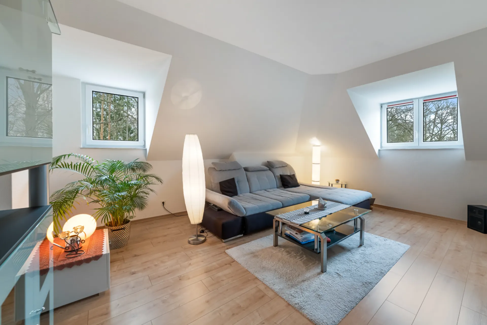 Wohnzimmer mit grauem Sofa, Glastisch, zwei Stehlampen und großen Fenstern mit Ausblick auf Bäume.