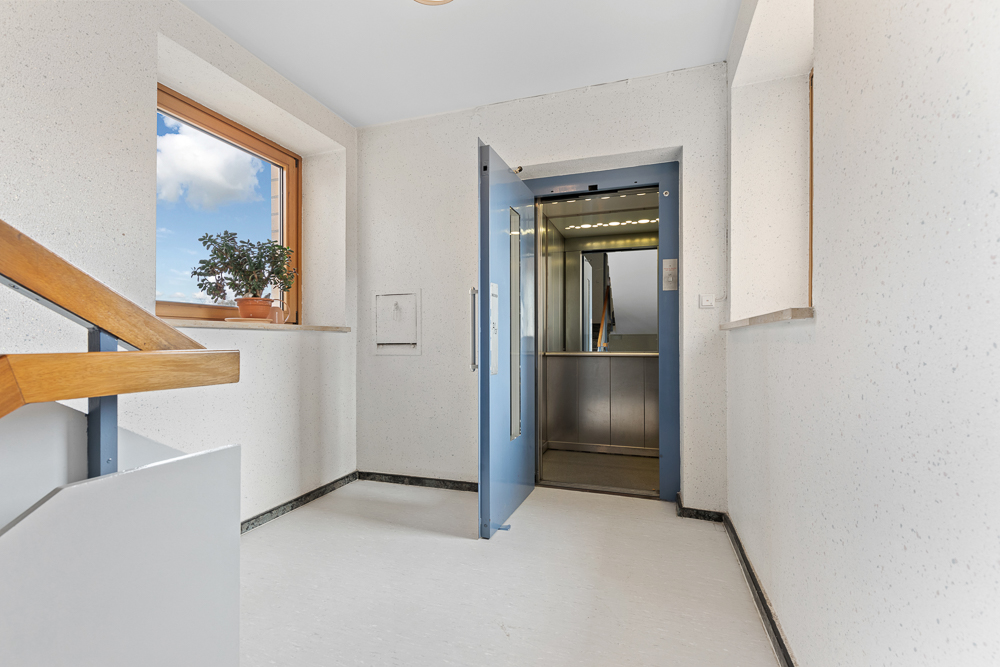Heller Flur mit offenem Aufzug, Fenster mit Blumentopf und Holzgeländer.