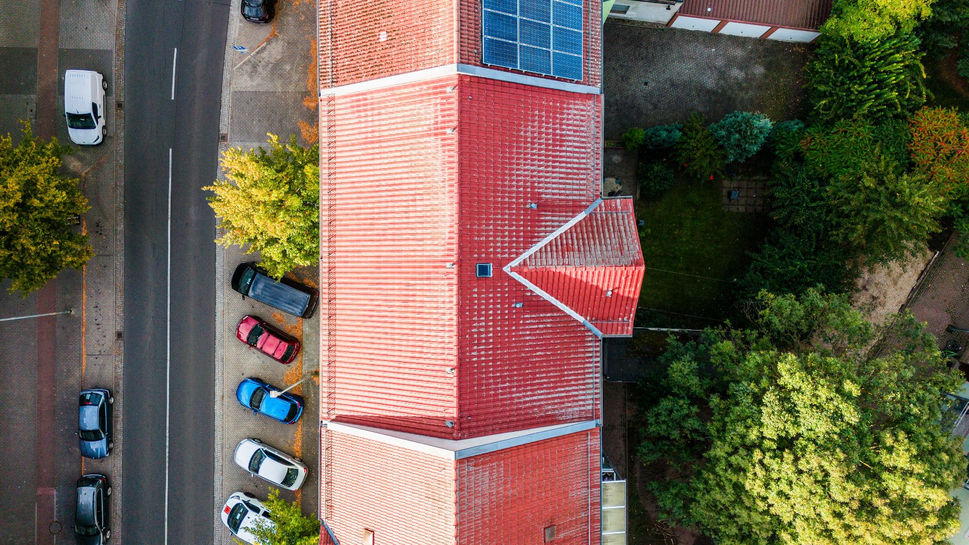 Draufsicht auf ein Haus mit rotem Dach und Solarpanelen, daneben Straße und geparkte Autos.