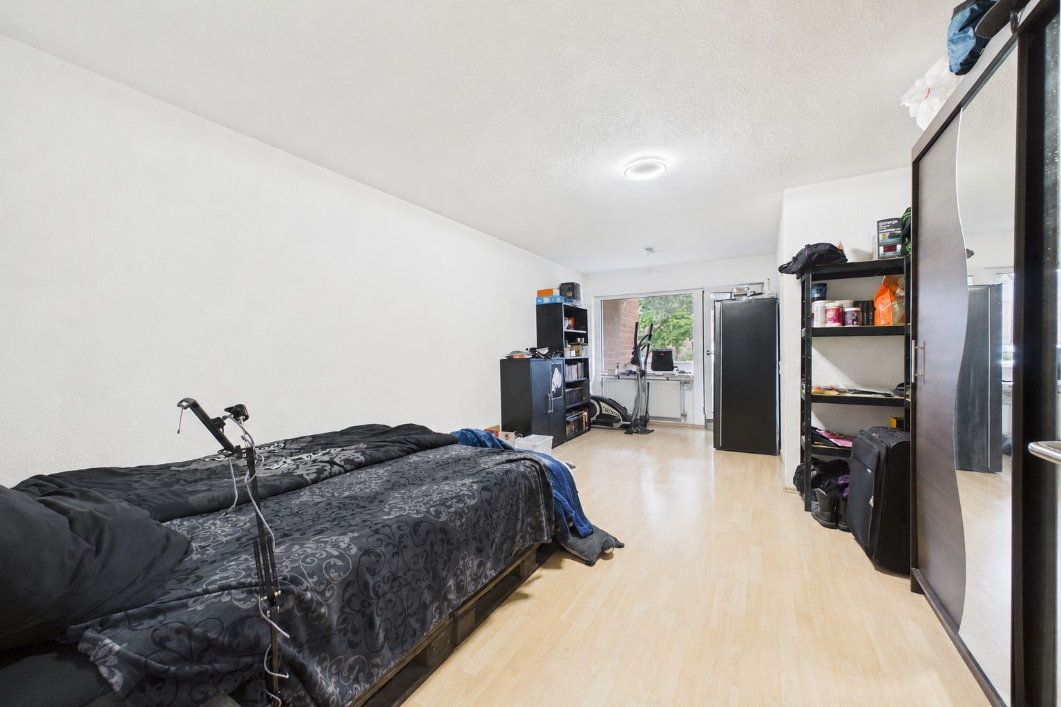 Schlafzimmer mit Einzelbett, Schrank, Regalen und Glastür zum Balkon.