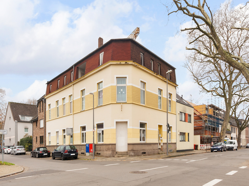 Eckhaus mit gelber und weißer Fassade, mehreren Fenstern und geparkten Autos an der Straße.