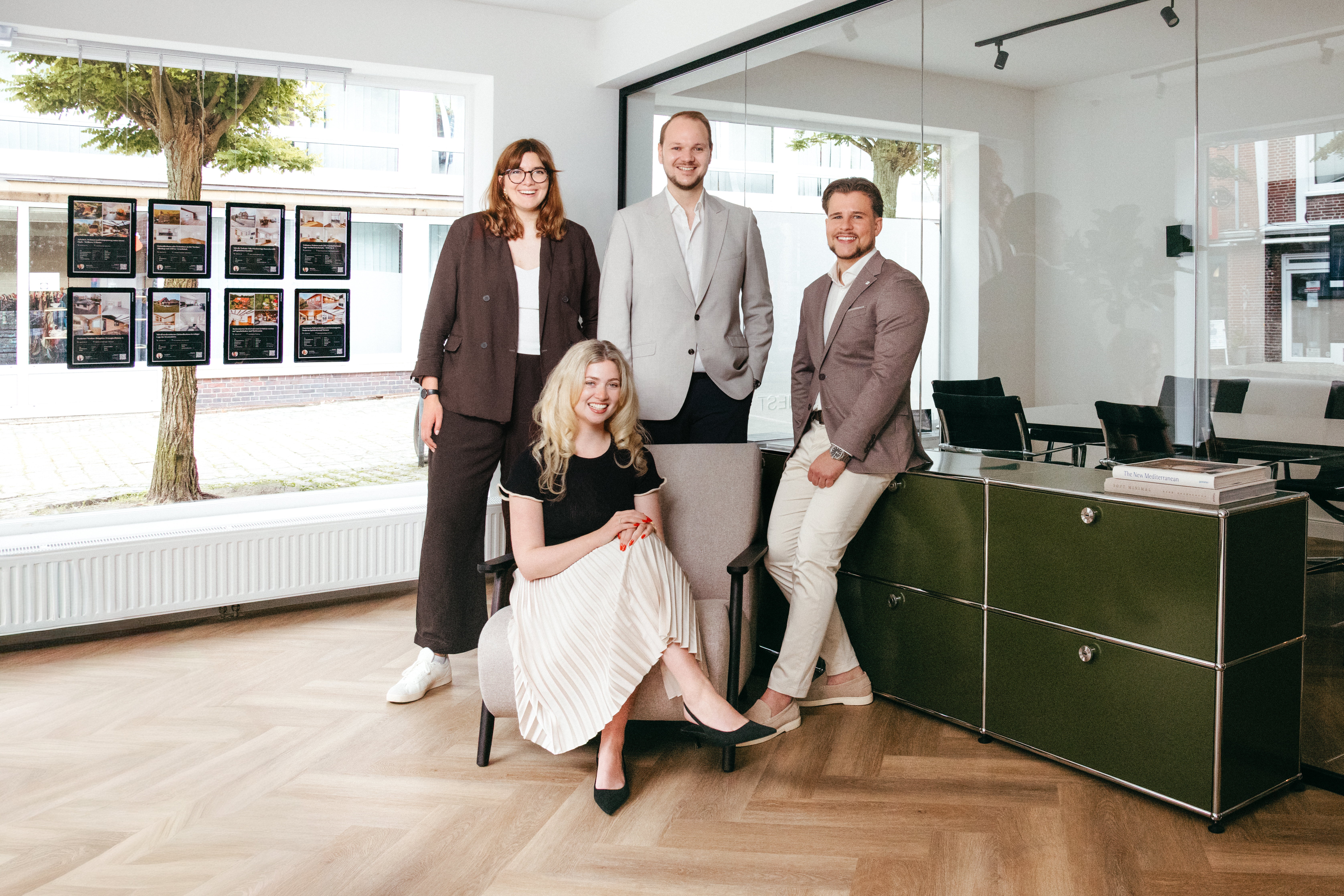 Vier Personen in Businesskleidung posieren in einem modernen Büro mit großen Fenstern und Holzboden.