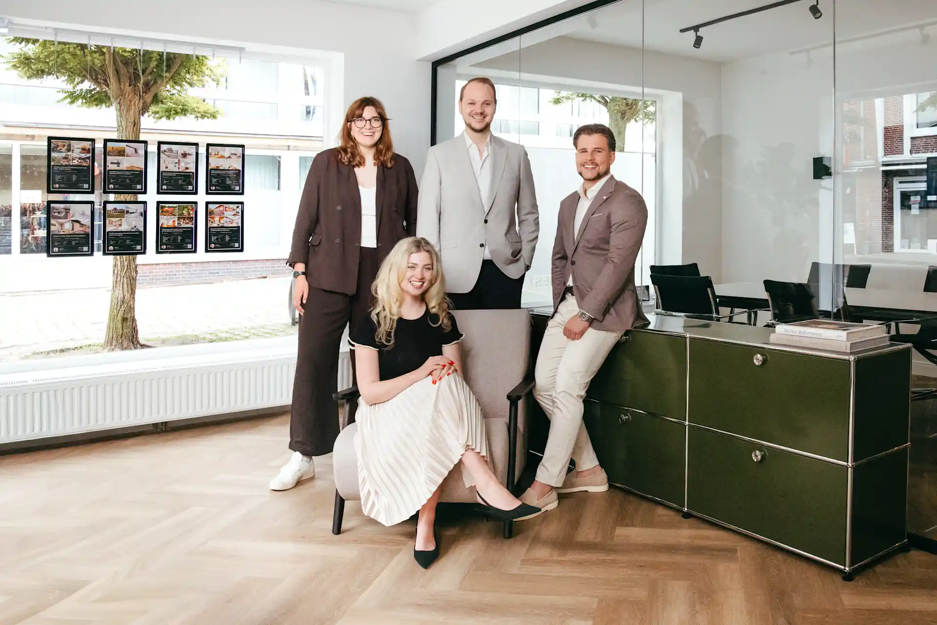 Vier Personen in Businesskleidung posieren in einem modernen Büro mit großen Fenstern und Holzboden.